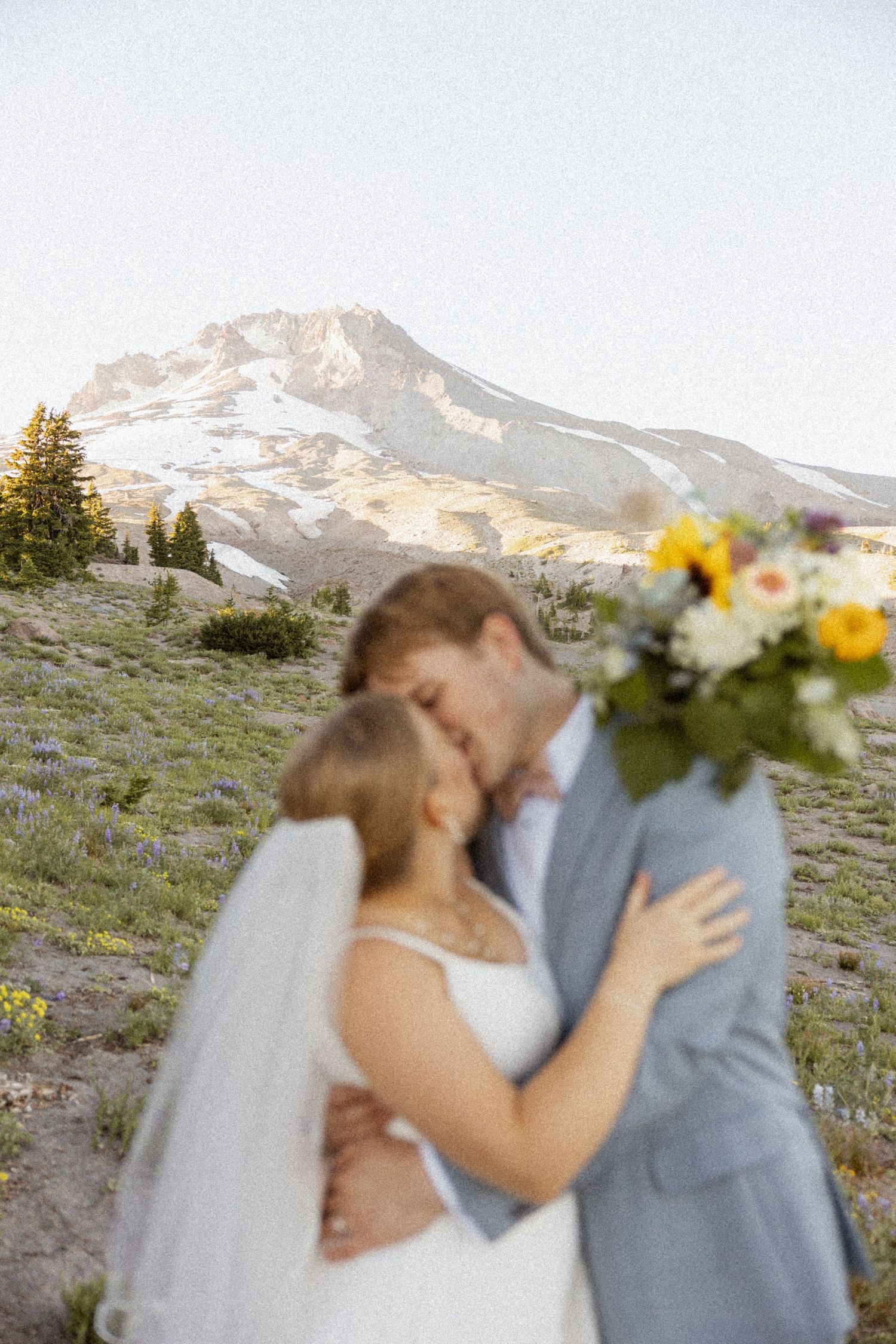 A wedding couple embraces against a mountain backdrop with yellow wildflowers in the foreground.
