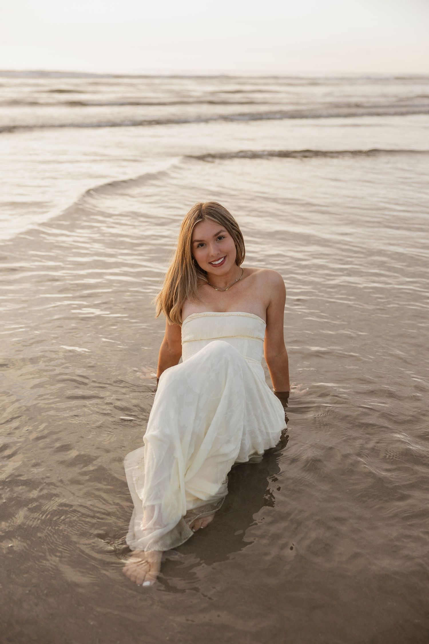 A woman in a flowing white dress wades through gentle ocean waves during a beach photoshoot at sunset.