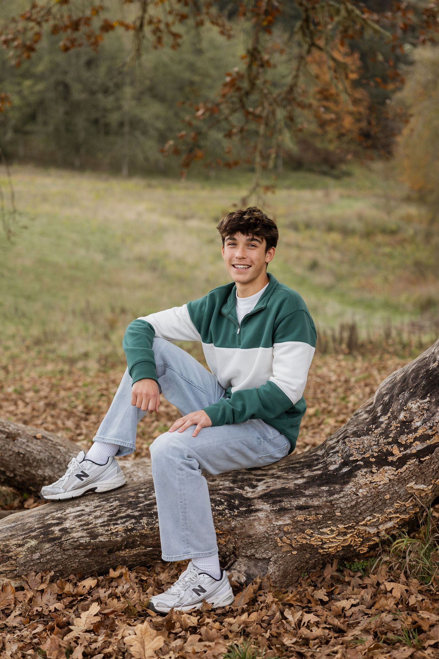 A person in casual wear sits on a fallen tree trunk surrounded by autumn leaves in an outdoor setting.