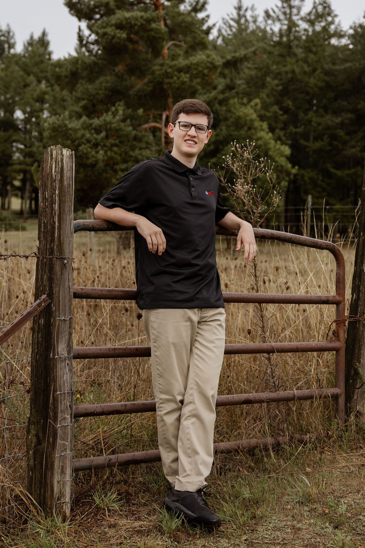 A casual outdoor portrait session with someone wearing a black polo and khaki pants leaning against a rustic wooden fence.