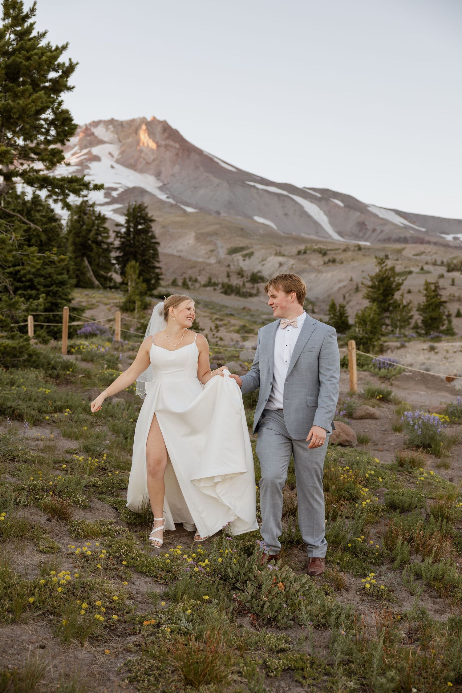 A wedding couple playfully walks through wildflowers with Mount Hood visible in the background.