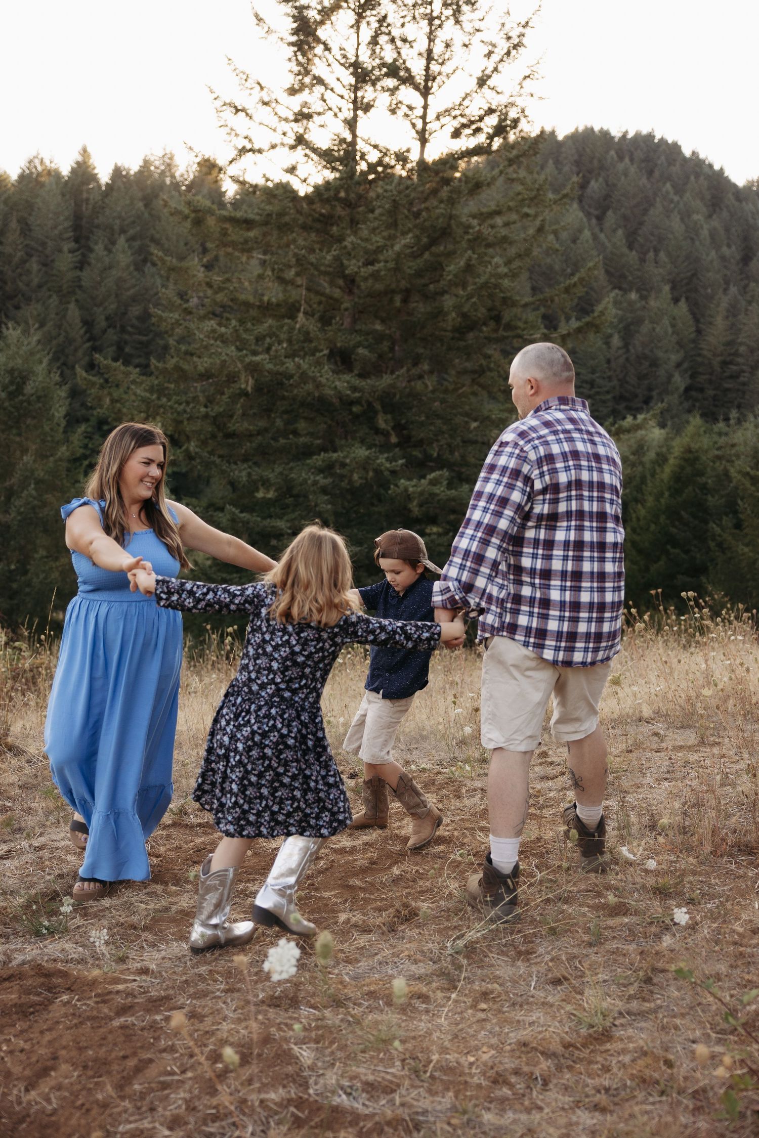 A family walks together holding hands through a grassy hillside at sunset with evergreen trees in the background.