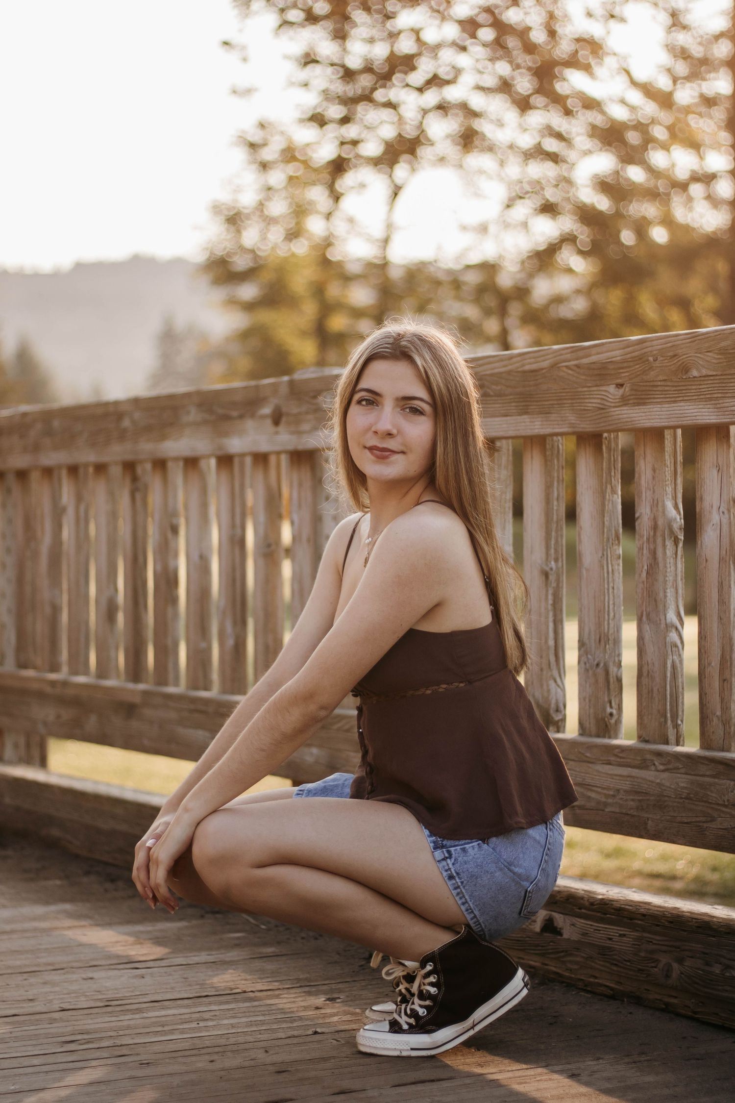 A person in denim shorts and a dark top poses casually on a wooden bridge deck at sunset.