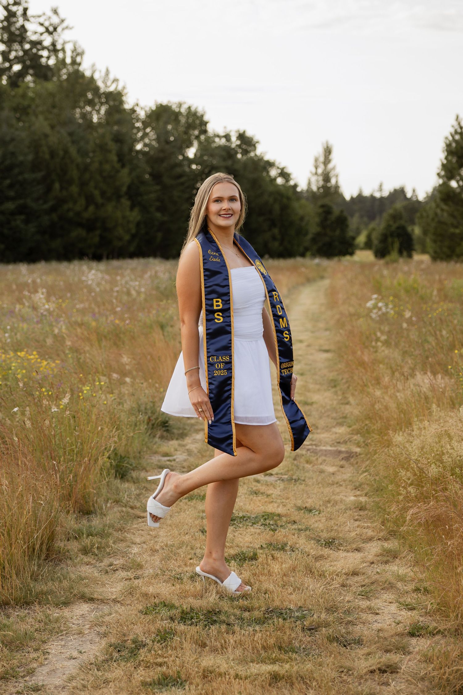 Graduate wearing blue sash poses in sunny field with wildflowers and dirt path during outdoor photo session.