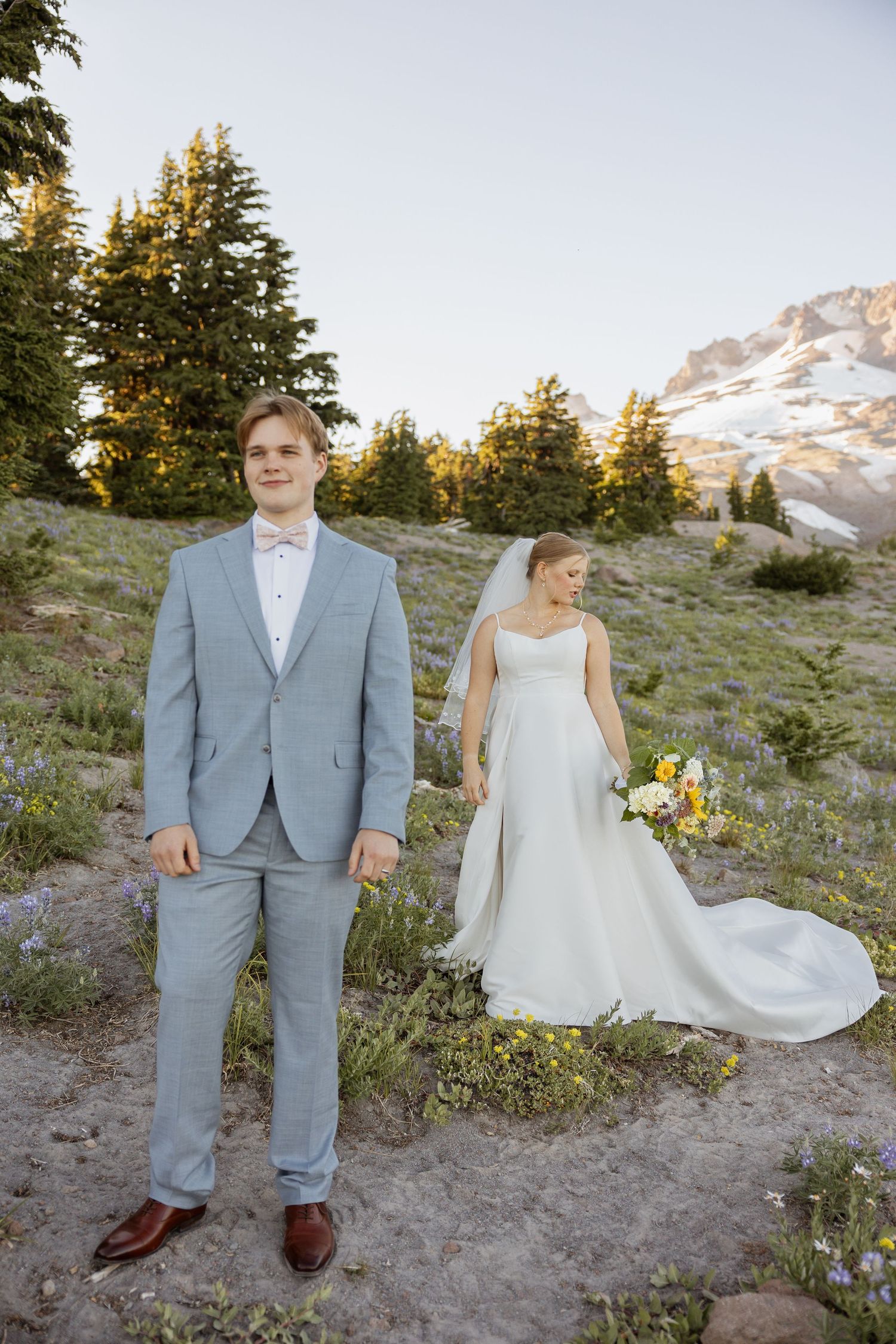 Wedding couple poses on mountainside with snow-capped peaks and evergreen trees in the background during golden hour.
