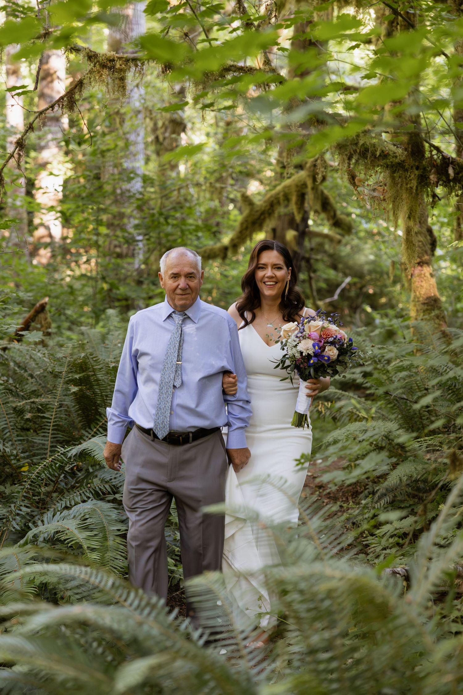 Two people walk arm in arm through a lush forest path surrounded by green ferns on their wedding day.