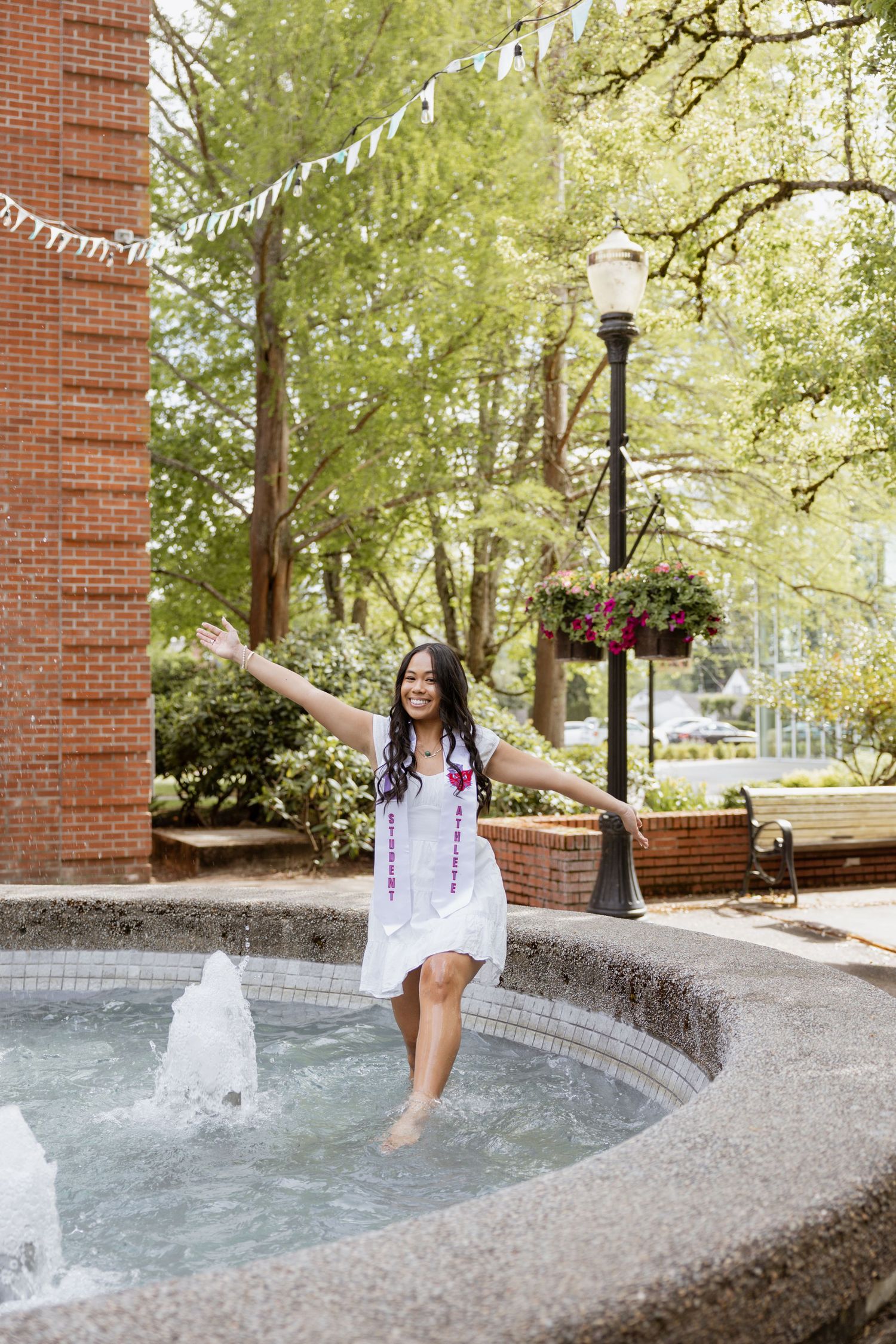 A person in a white dress sits joyfully at the edge of a curved fountain with water features on a sunny spring day.
