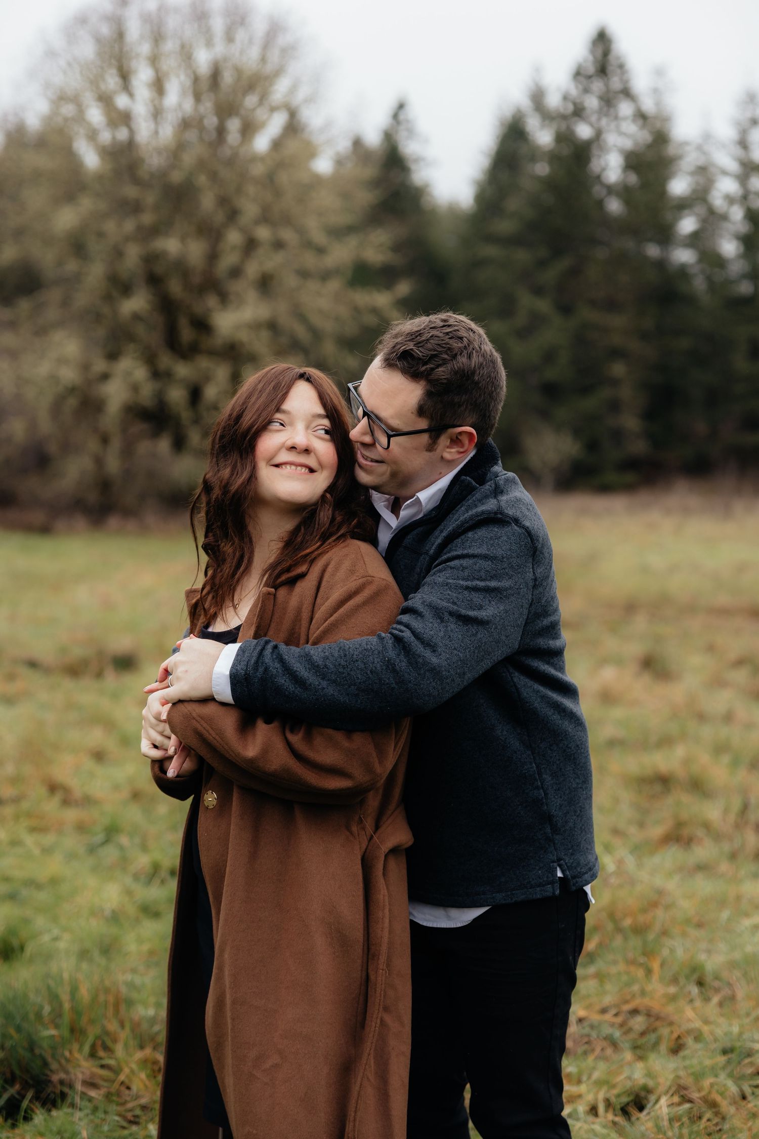 A couple embraces lovingly in a field with evergreen trees in the background during an outdoor photo session.