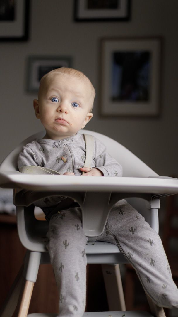 Baby in grey onesie sitting in white high chair making playful expressions.