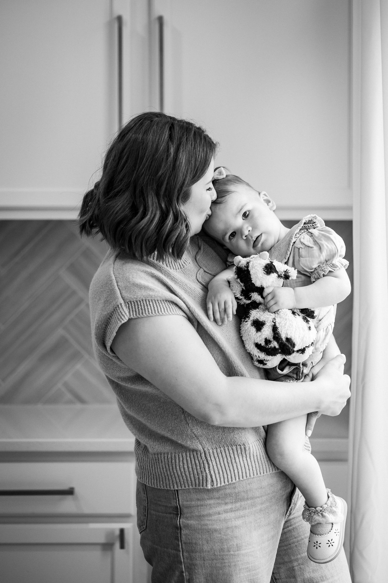 Black and white photo of a mother tenderly holding and kissing her baby girl in a bright kitchen.