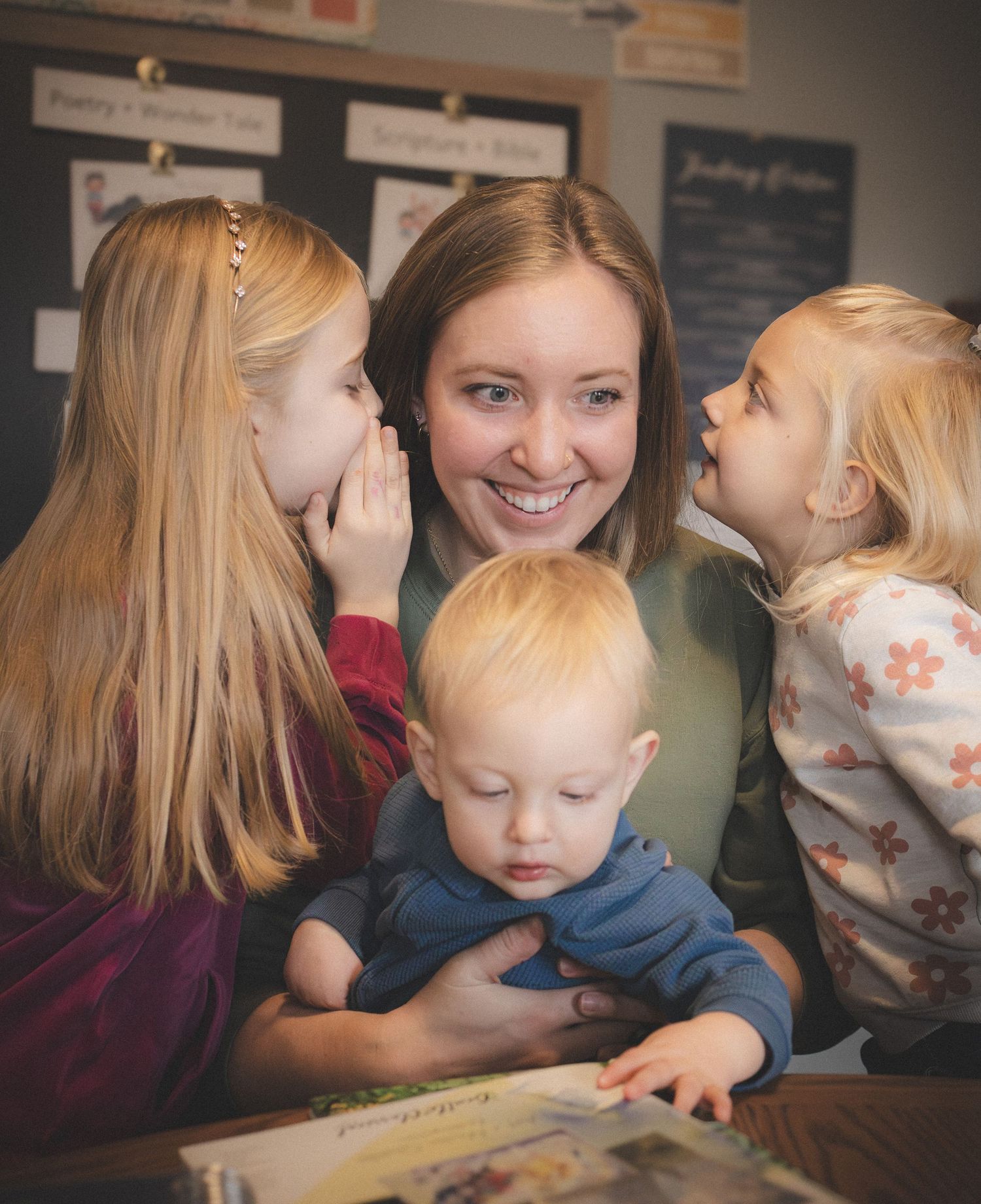 A woman with long blonde hair is smiling and interacting with two young children in a classroom setting.