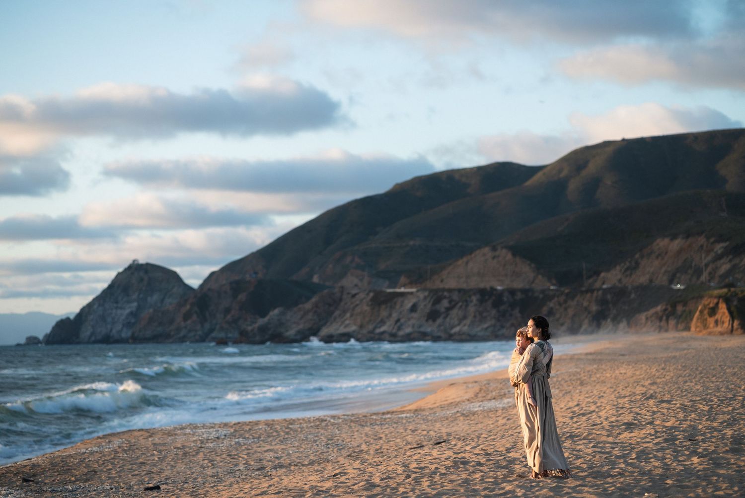 Couple embracing on a sandy beach at golden hour with dramatic coastal cliffs and ocean waves in the background.