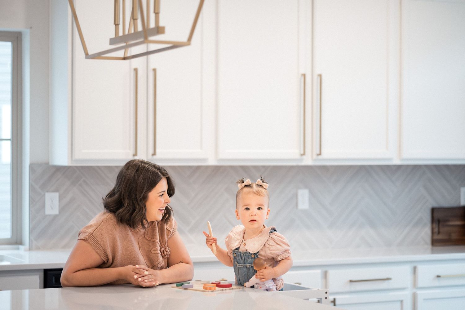 A mother and baby girl sit together at a white kitchen island in a bright modern kitchen.