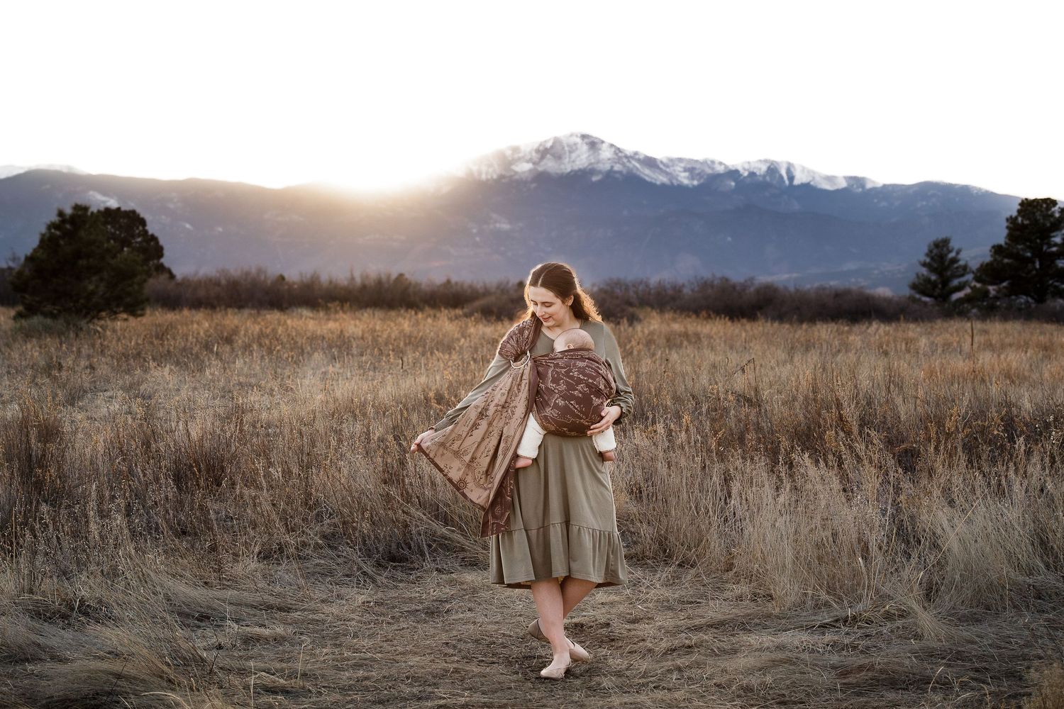 Woman in bohemian dress holding floral shawl in golden field with snow-capped mountains at sunset.