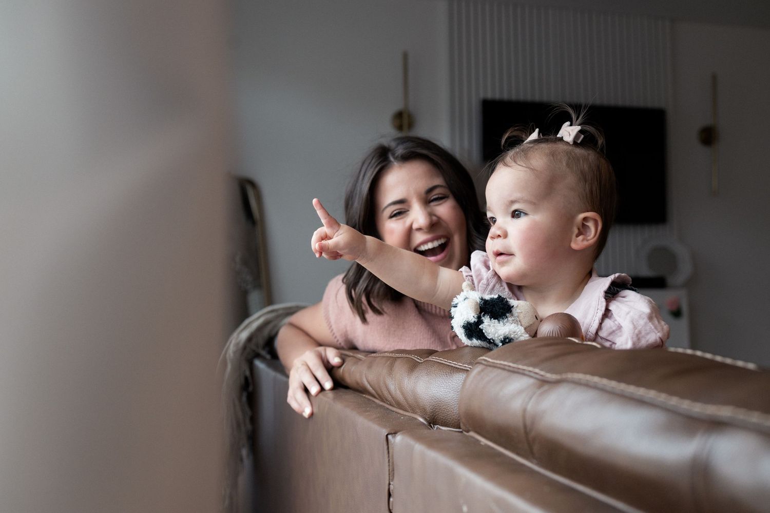 A smiling woman points at something while holding a baby girl in a pink dress on a leather sofa indoors.