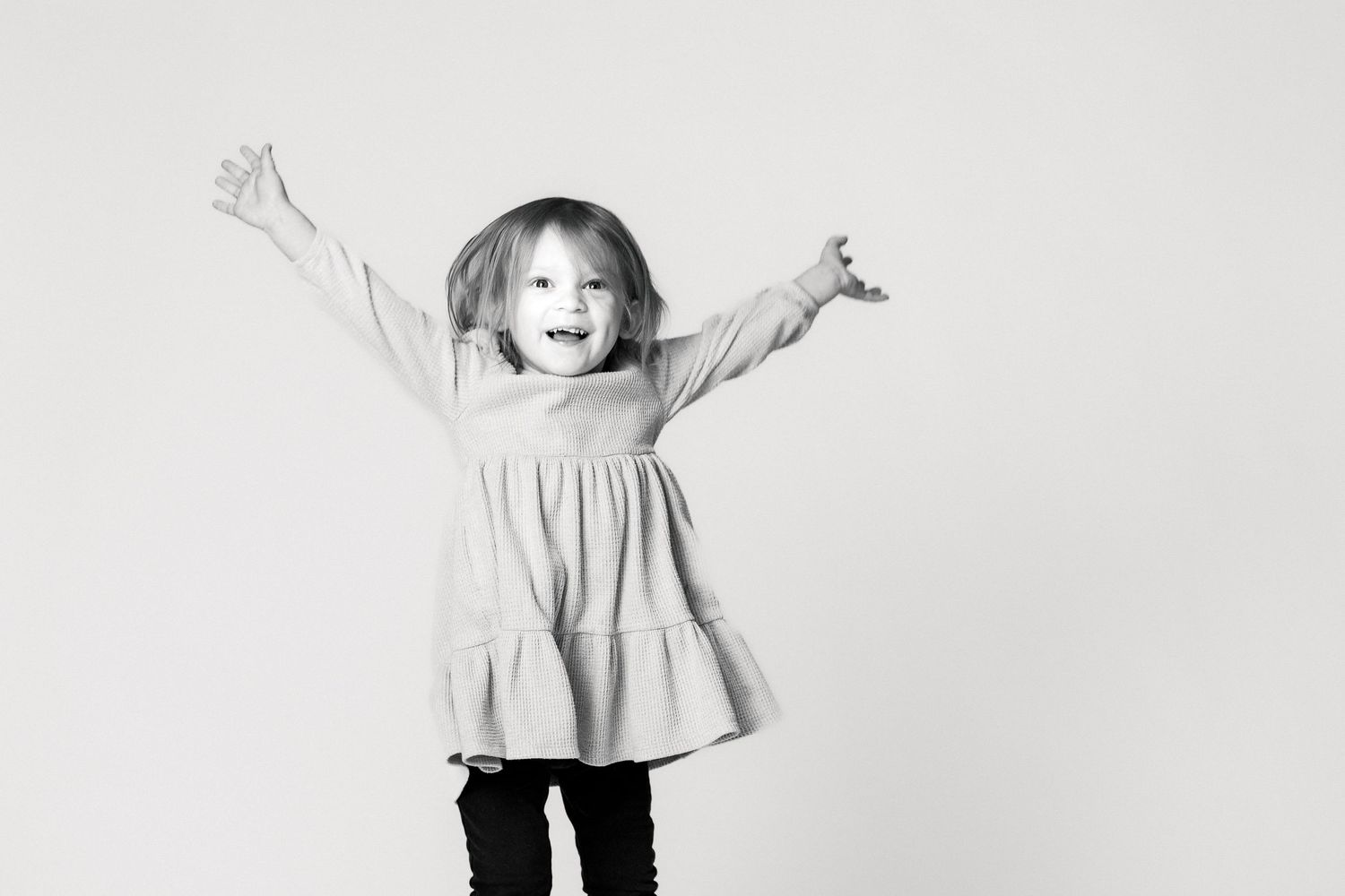 A young girl with a joyful expression stands with her arms raised, wearing a dress and smiling happily.