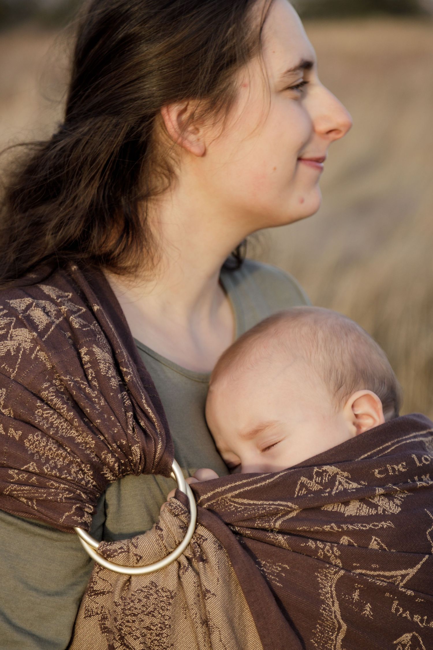 A young mother carries her sleeping newborn in a brown Tolkien-themed ring sling outdoors in a golden field.