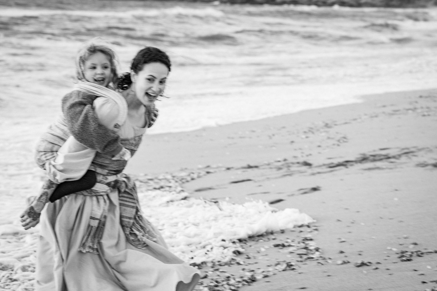 Black and white photo of a laughing woman carrying a child on her back while running along a sandy beach.