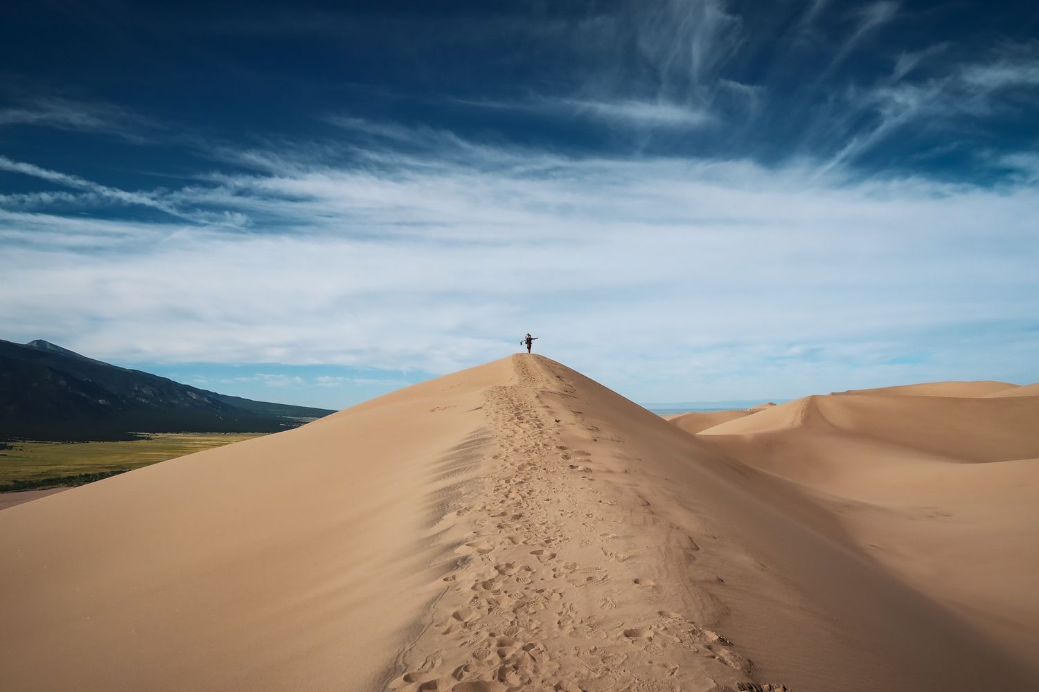 A distant figure stands atop a massive sand dune in Great Sand Dunes National Park, Colorado against a cloudy blue sky.
