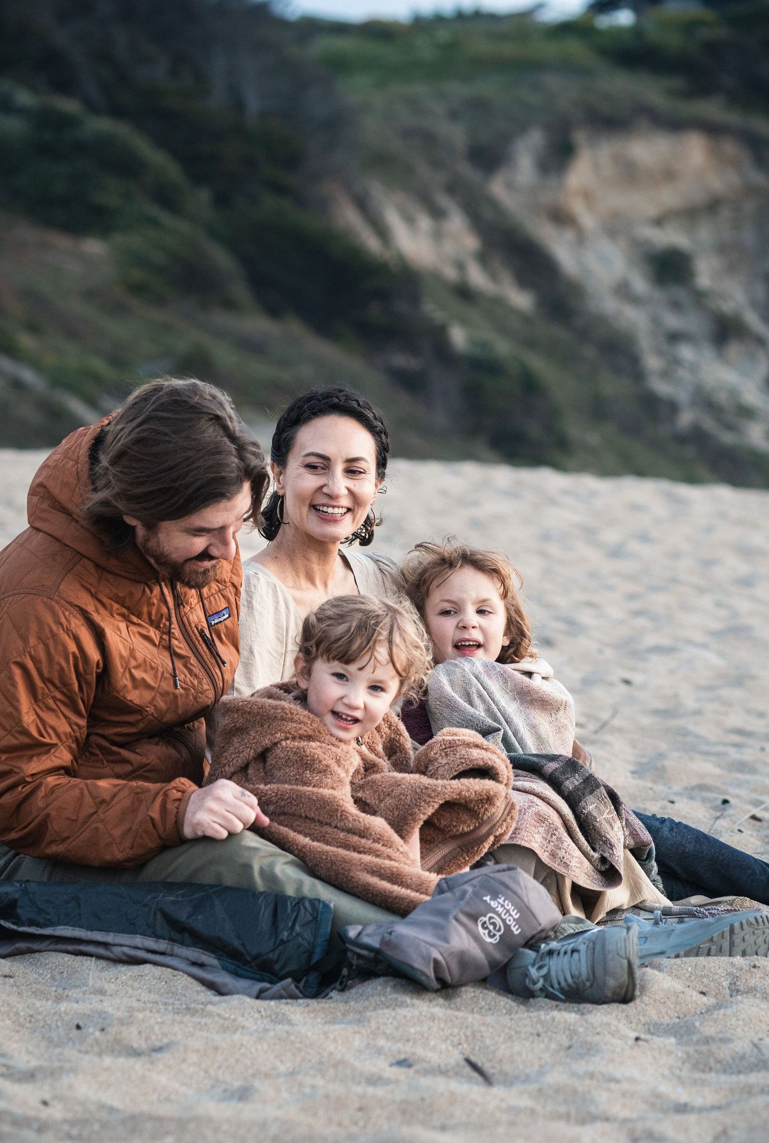Family of four wrapped in cozy blankets sitting together on a sandy beach with cliffs in the background.