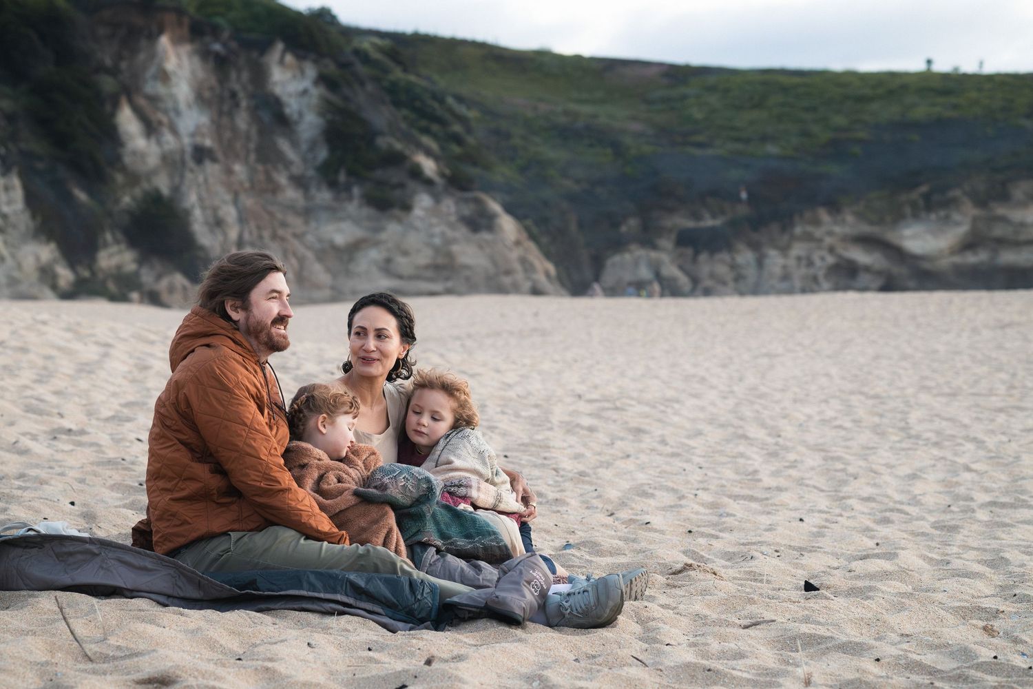 Family of four sitting together on a sandy beach, smiling warmly, with rocky cliffs in the background.