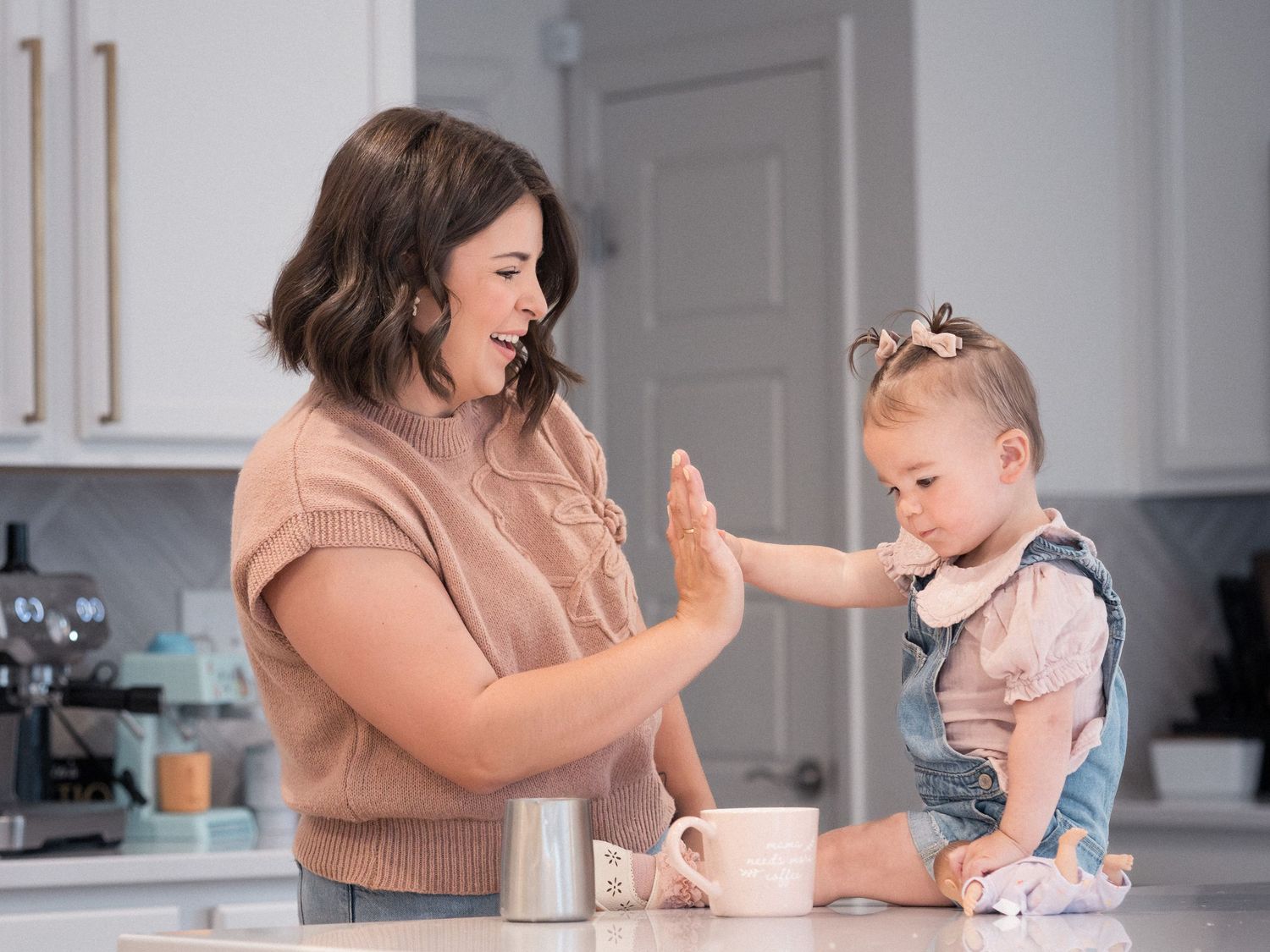 A mom and toddler girl share a high five at a kitchen counter, smiling together in a bright, modern kitchen.