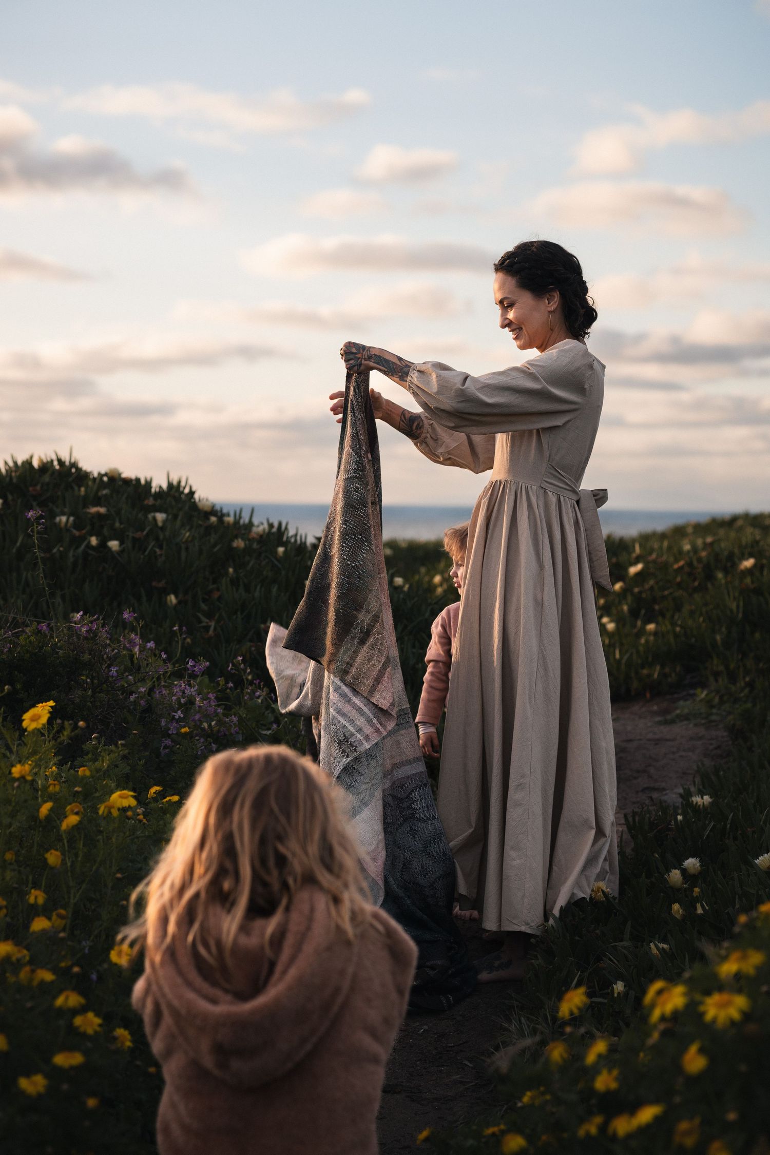 Woman in linen dress holding blanket with two children among wildflowers near ocean at sunset.