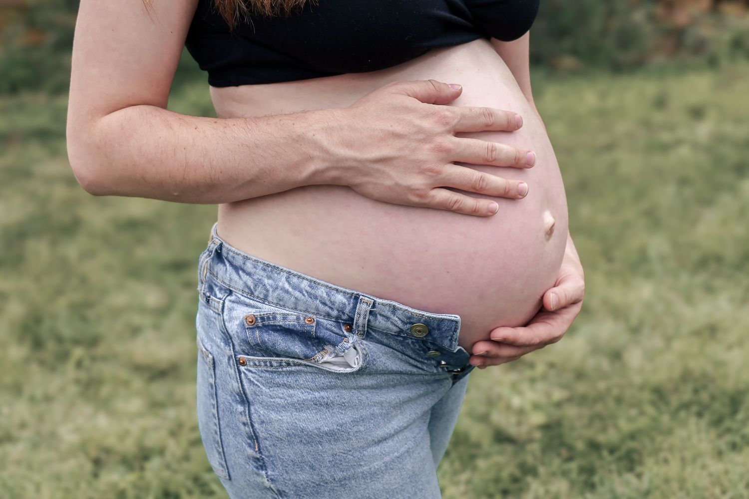 Close up view of pregnant belly with hands gently cradling the baby bump while wearing denim jeans.