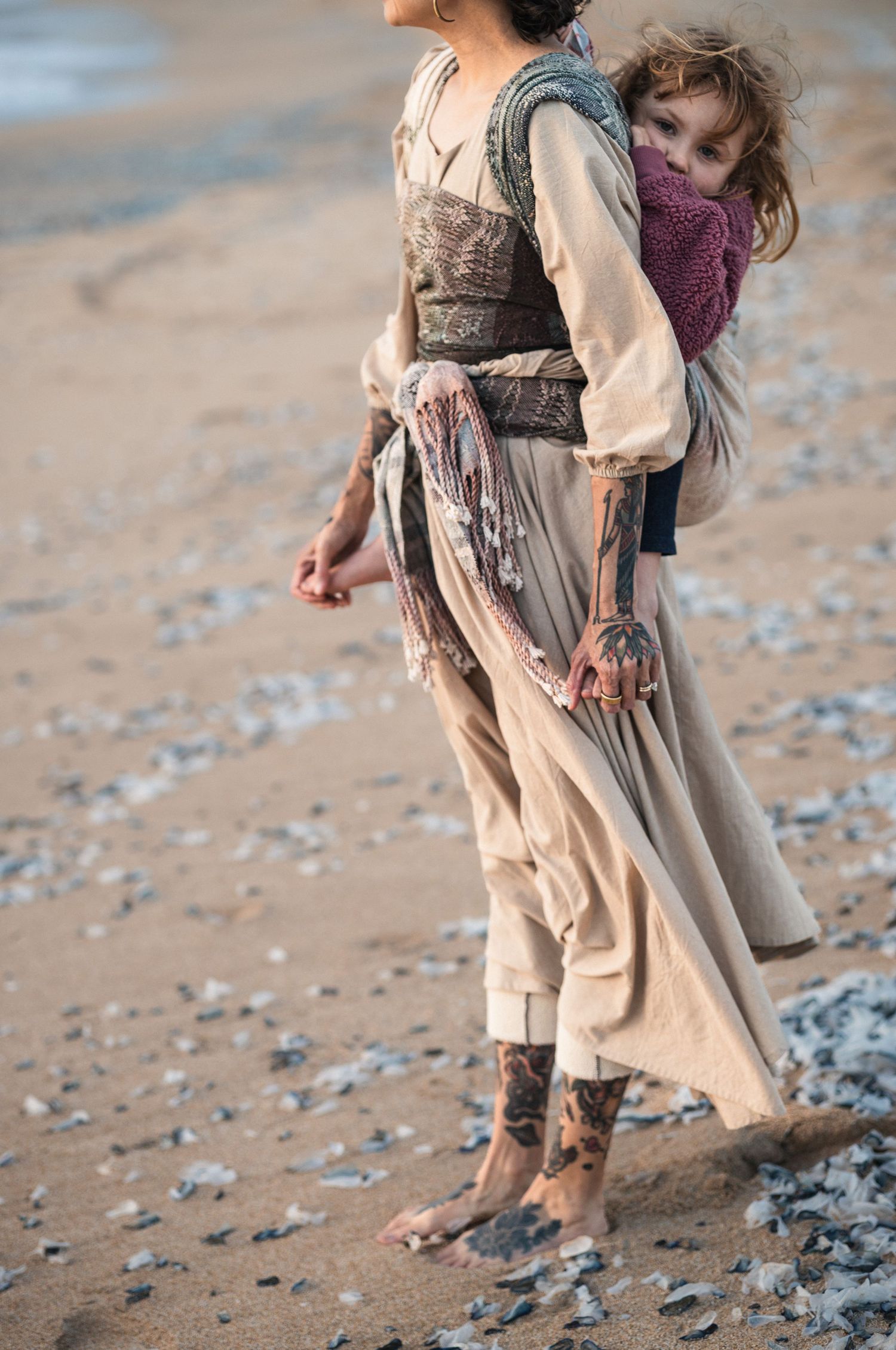 Tattooed woman in beige dress carries child on beach, barefoot on sandy shore.