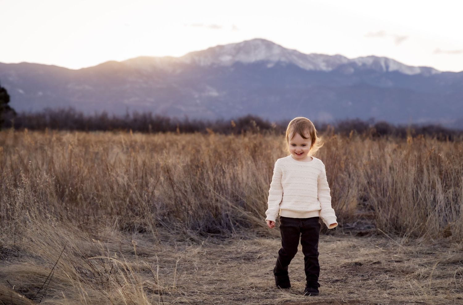 A young child stands in a grassy field, with mountains visible in the background on a hazy day.