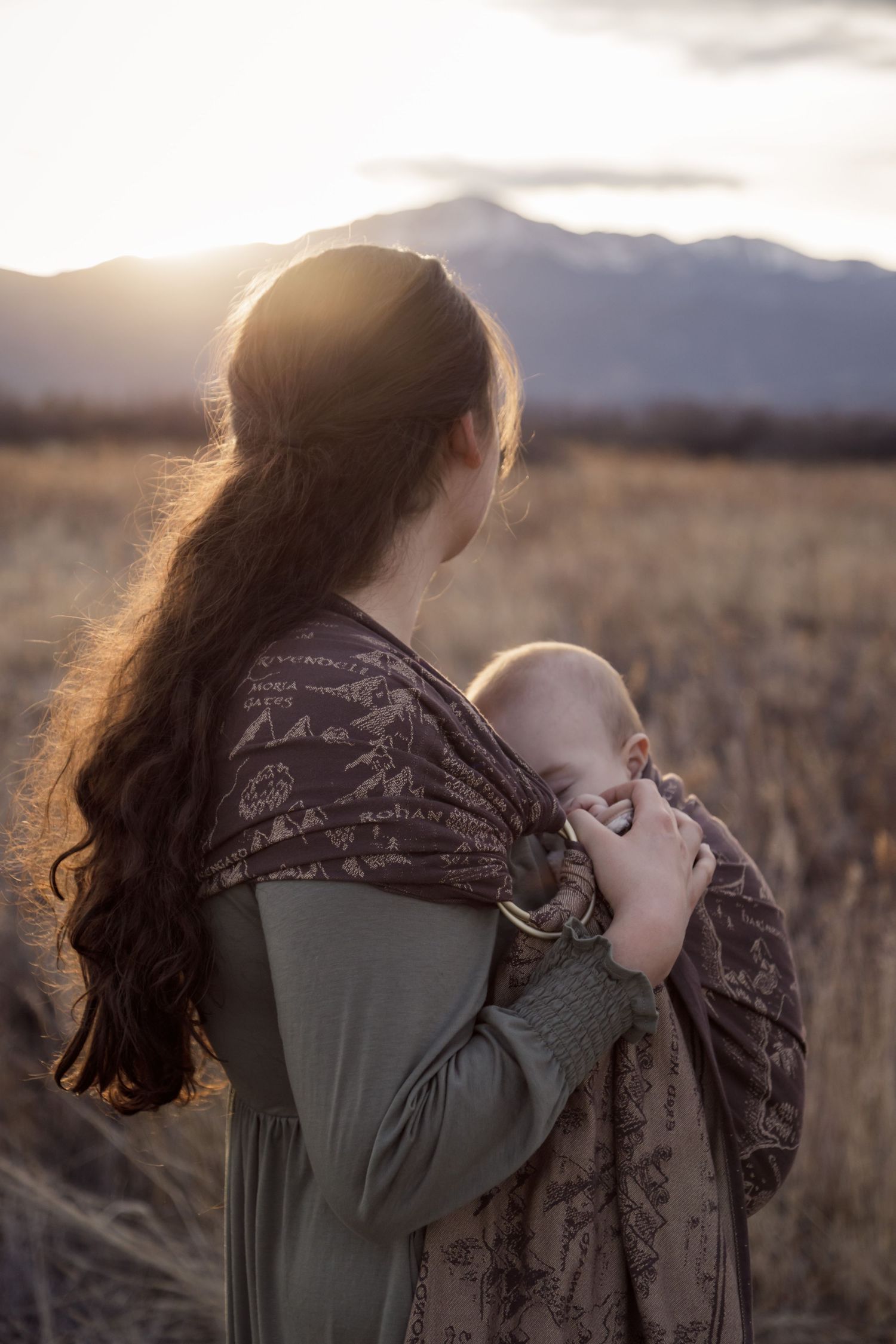 A silhouetted figure with long hair holds a baby in a wrap carrier against a sunset mountain backdrop in a grassy field.