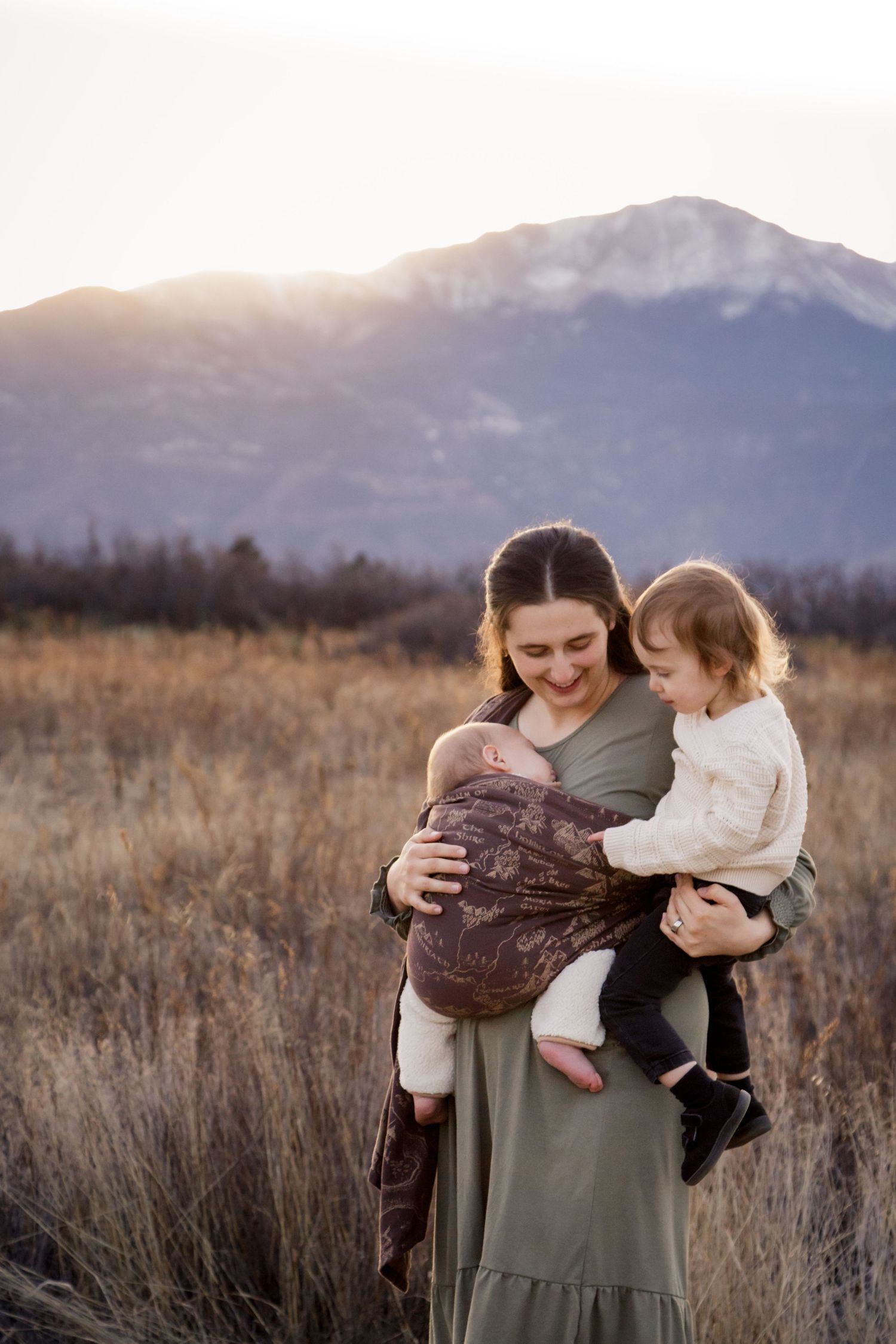 Mother holding newborn and toddler in a sunlit mountain meadow, wearing a green dress, smiling warmly outdoors.
