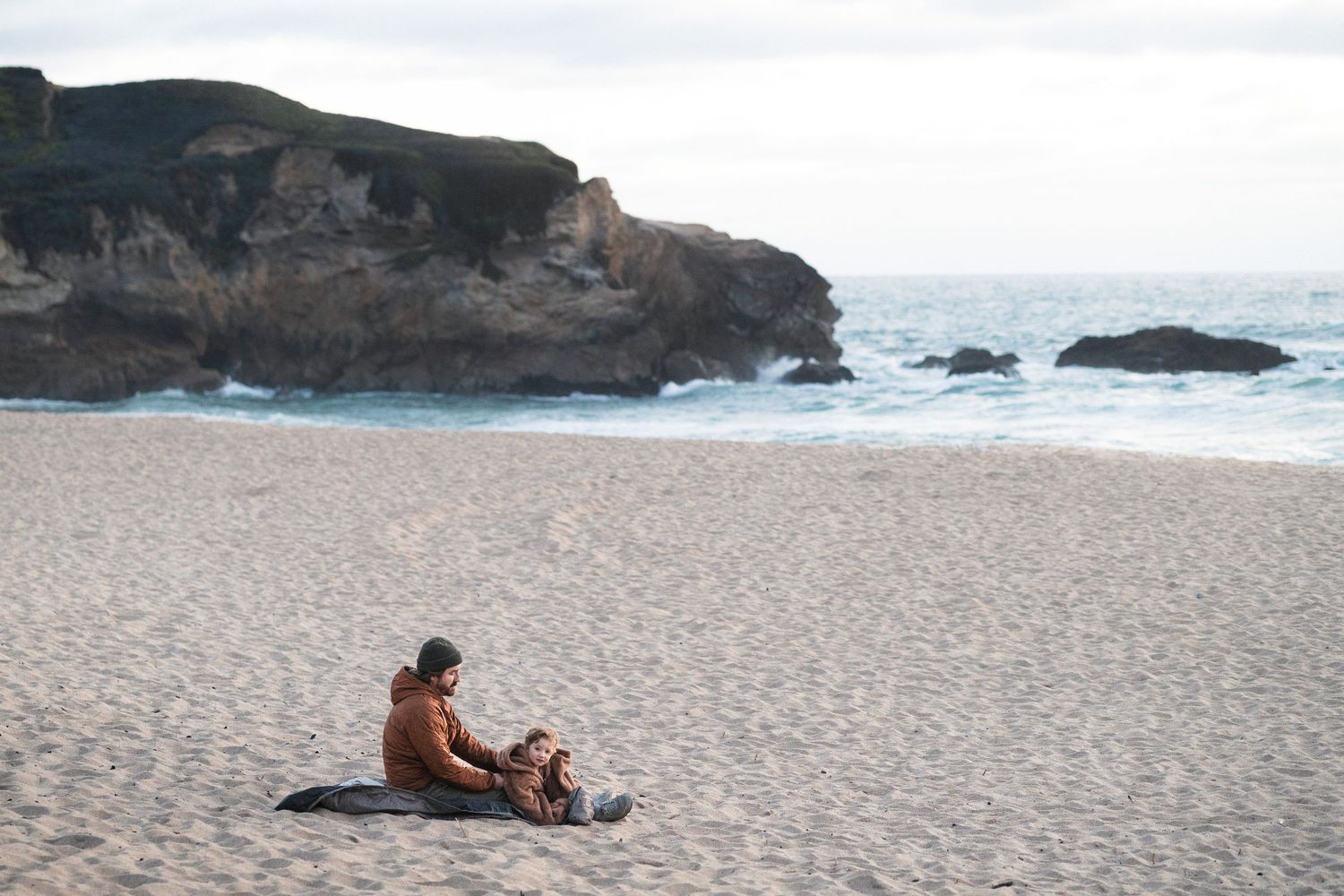 Two people sitting on a blanket on a sandy beach with rocky cliffs and ocean waves in the background.