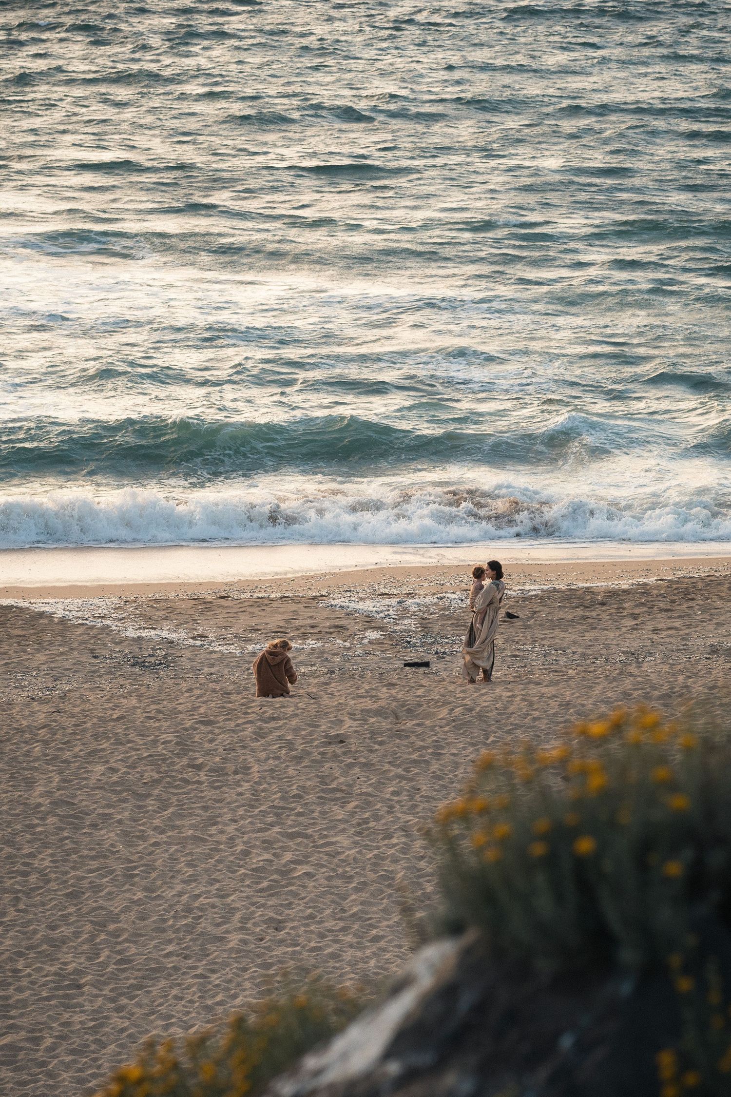 Two people and a dog enjoy a peaceful sunset walk on a sandy beach with gentle ocean waves in the background.