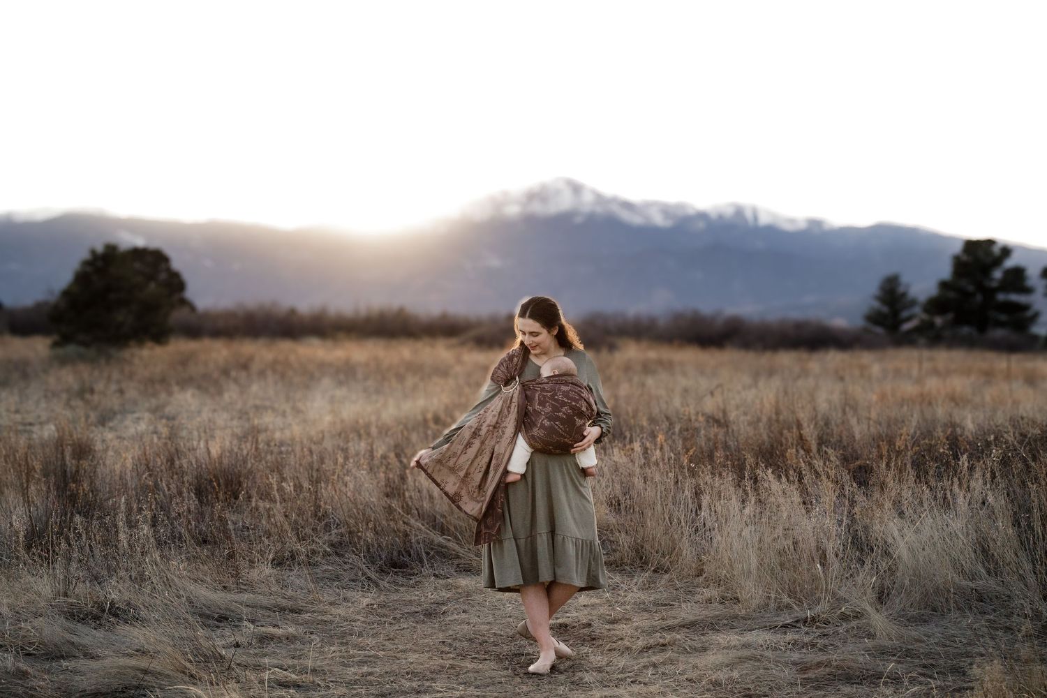 Woman in green skirt and brown shawl walks through golden field at sunset with snow-capped mountains behind her.