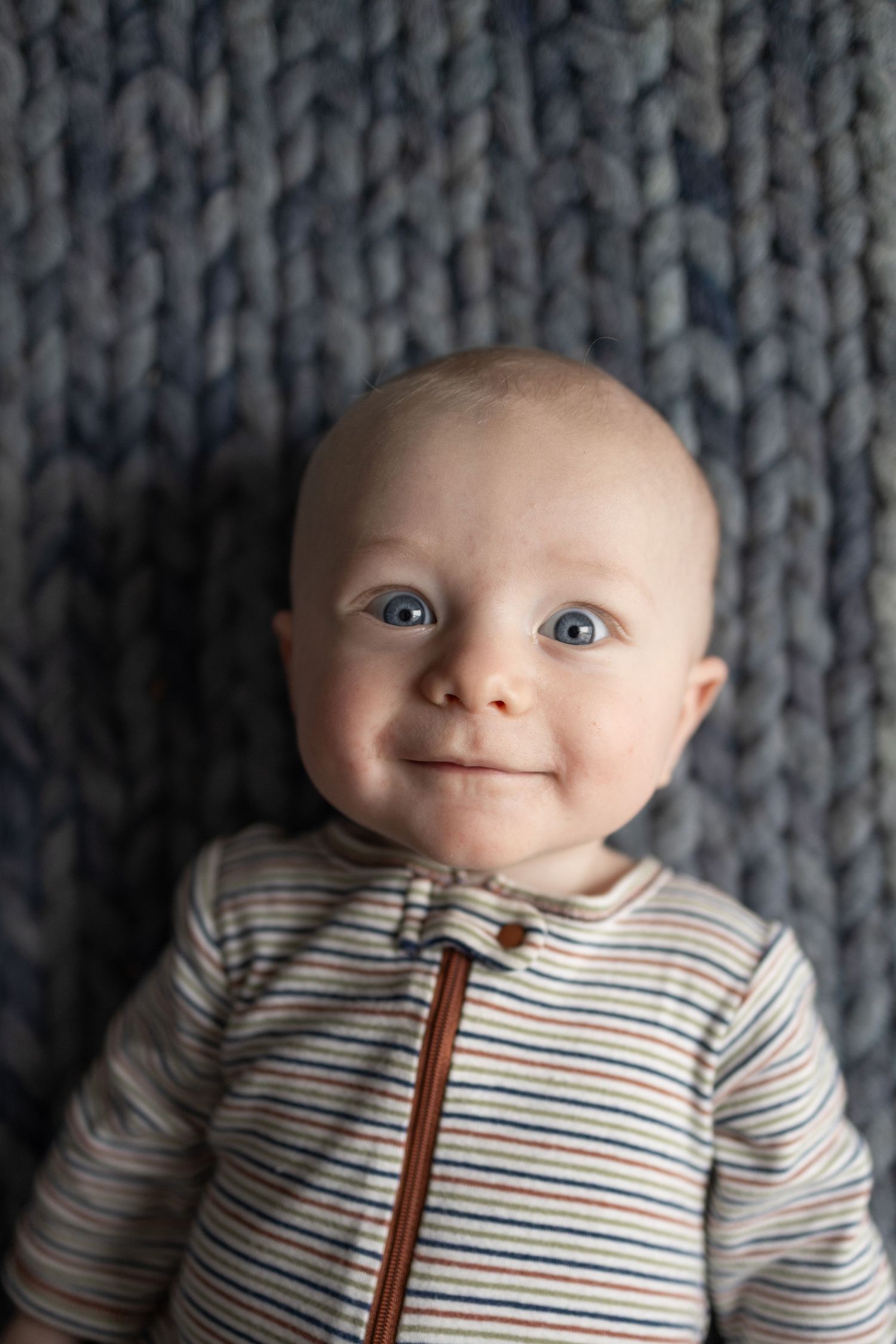 A baby in a striped onesie shows a range of adorable expressions while lying on a gray knit blanket.