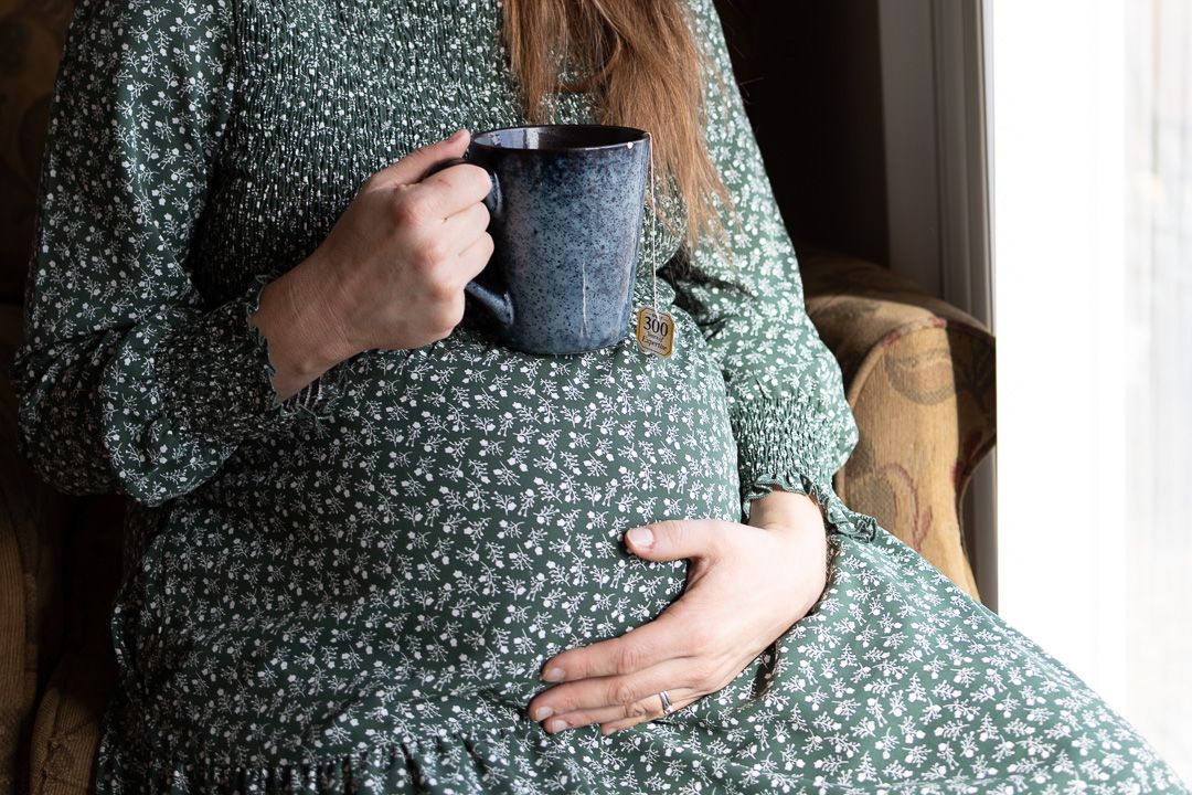 Person in floral dress holding blue mug while sitting by window during pregnancy.