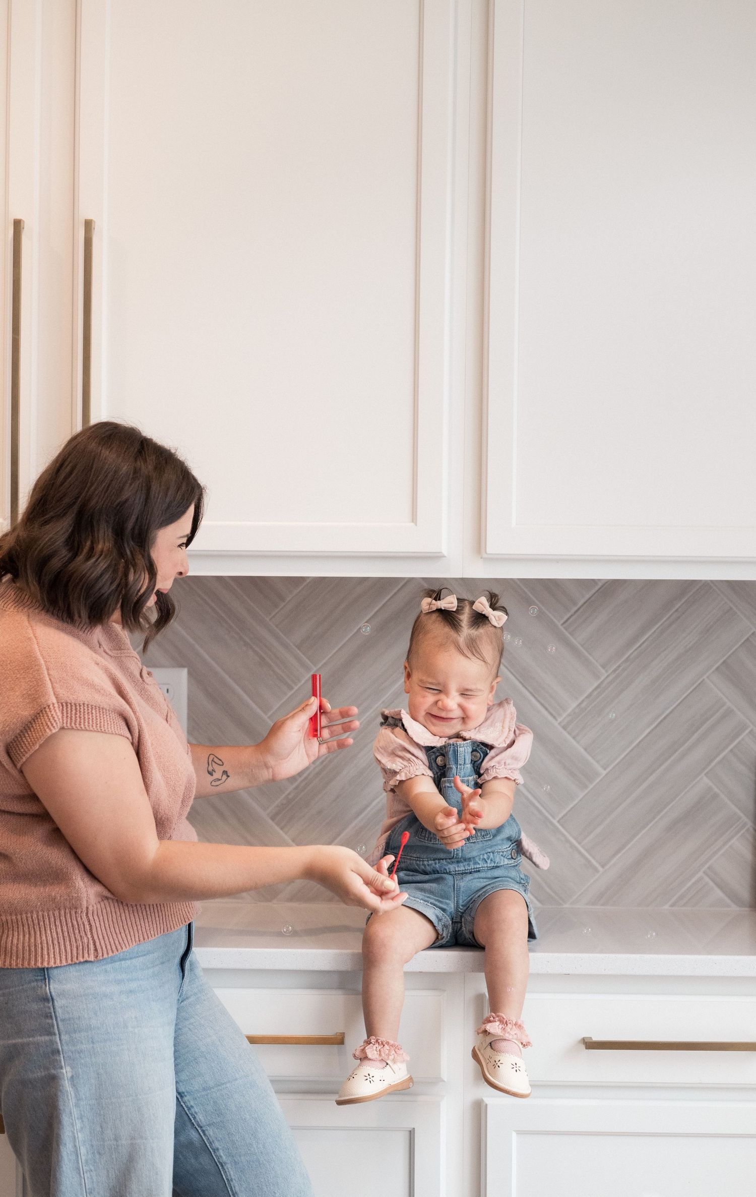 A woman holds a red toy while a smiling baby in denim overalls sits on a white kitchen counter.