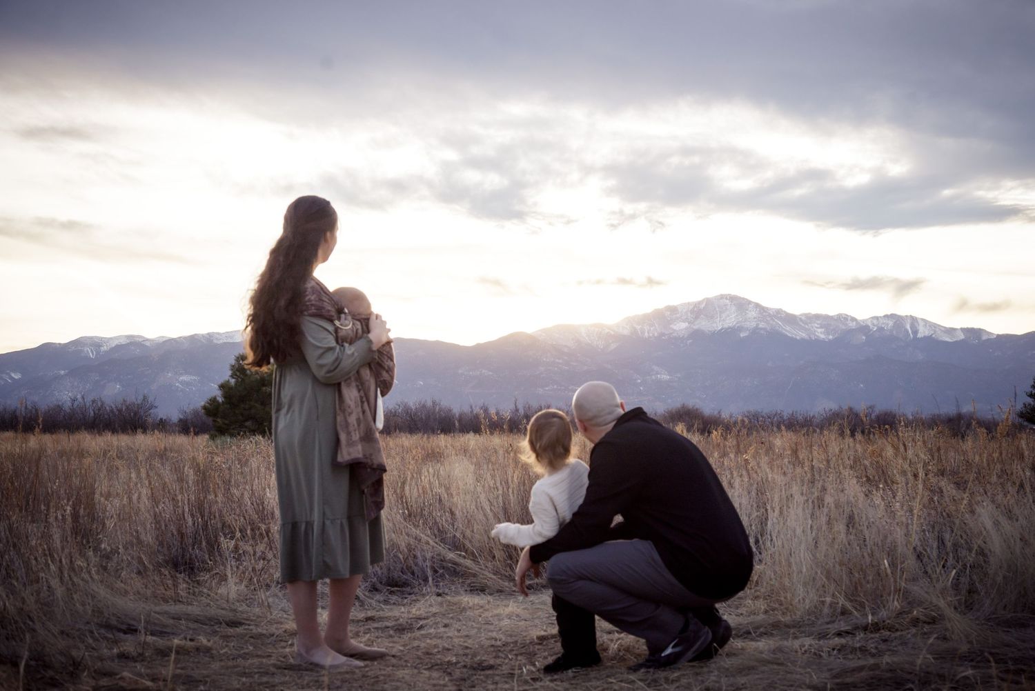 A man kneels in a field as a woman stands holding a child, with mountains in the background.