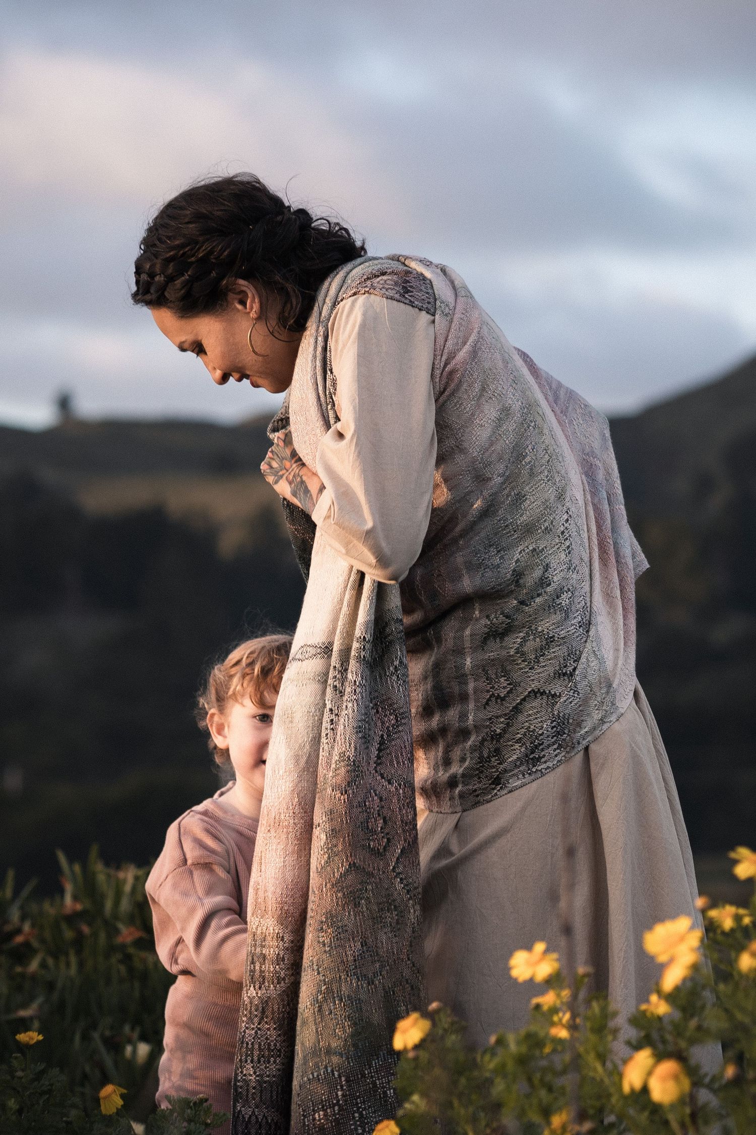 A woman in traditional dress tenderly looks down at a small child among yellow wildflowers at dusk.