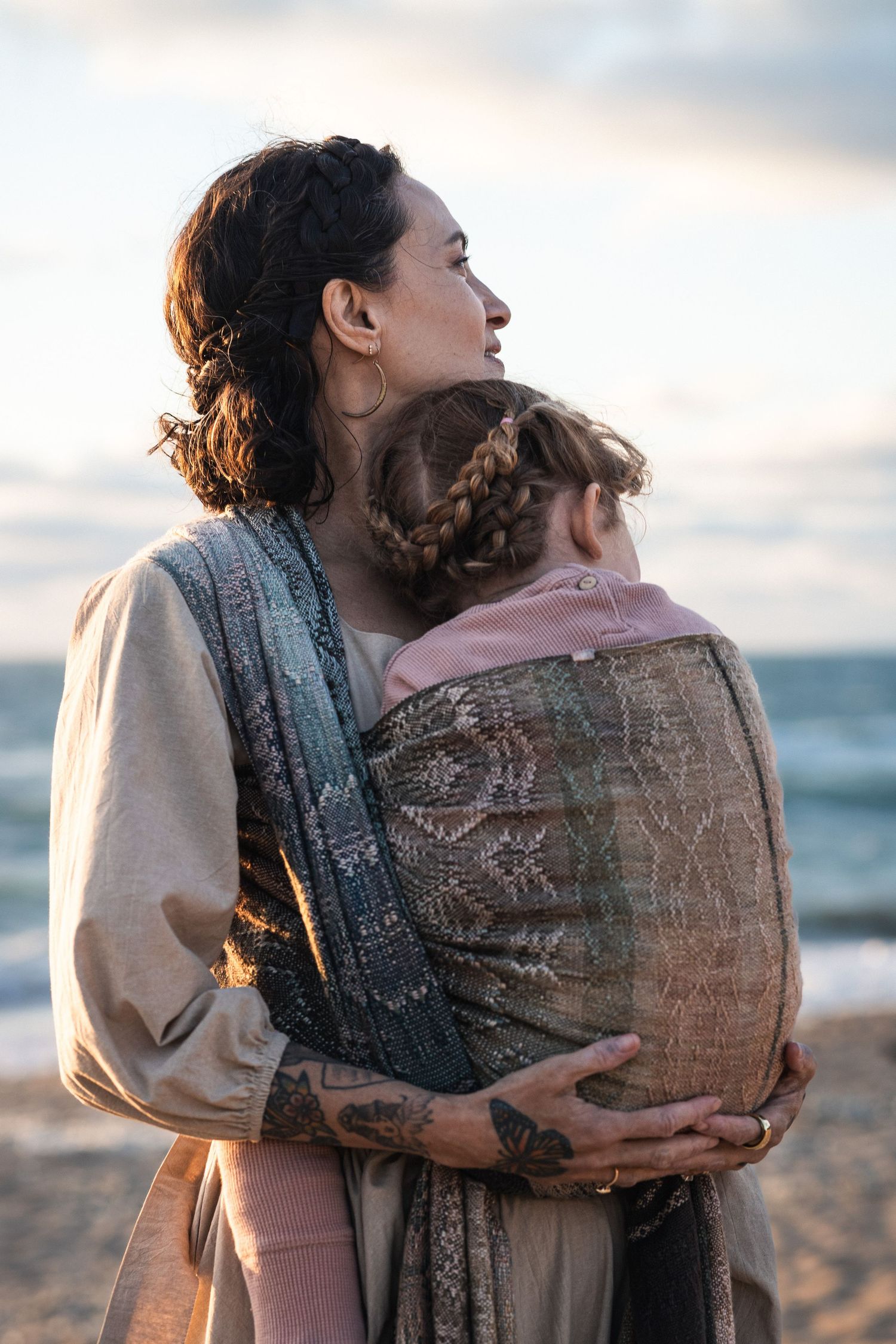 Mother carrying child in a woven baby wrap carrier on a beach at sunset, both with braided hair.