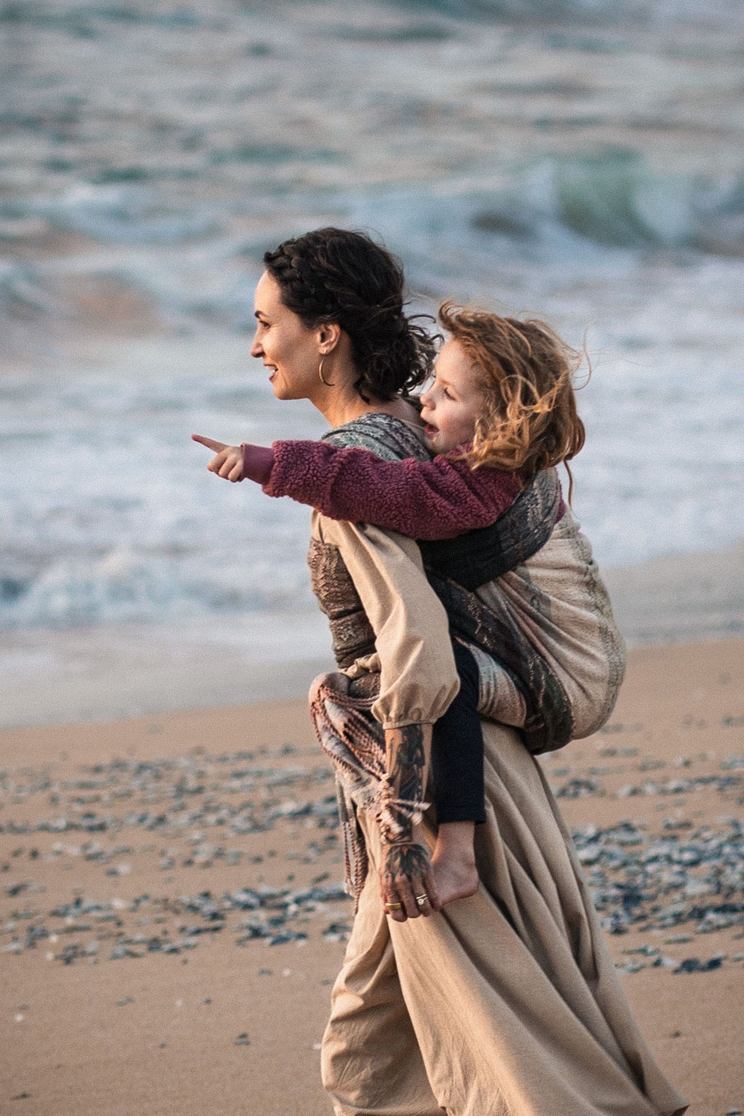 A woman carries a child on her back along a sandy beach, with ocean waves in the background.