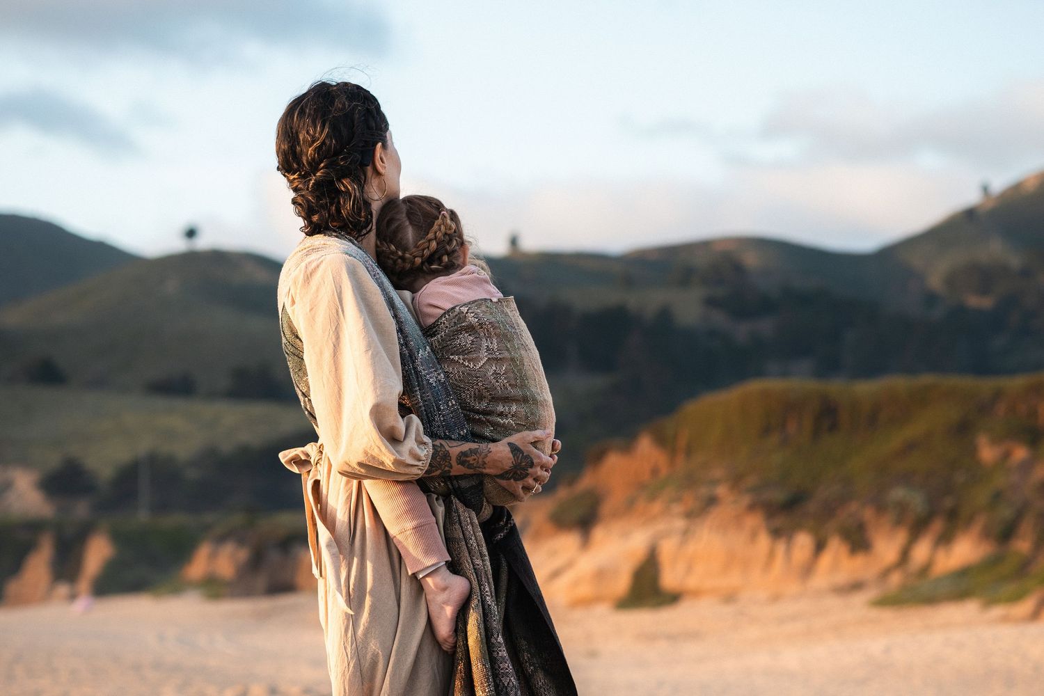 Woman carrying baby in carrier on sandy beach with green hills and dramatic cliffs in background.