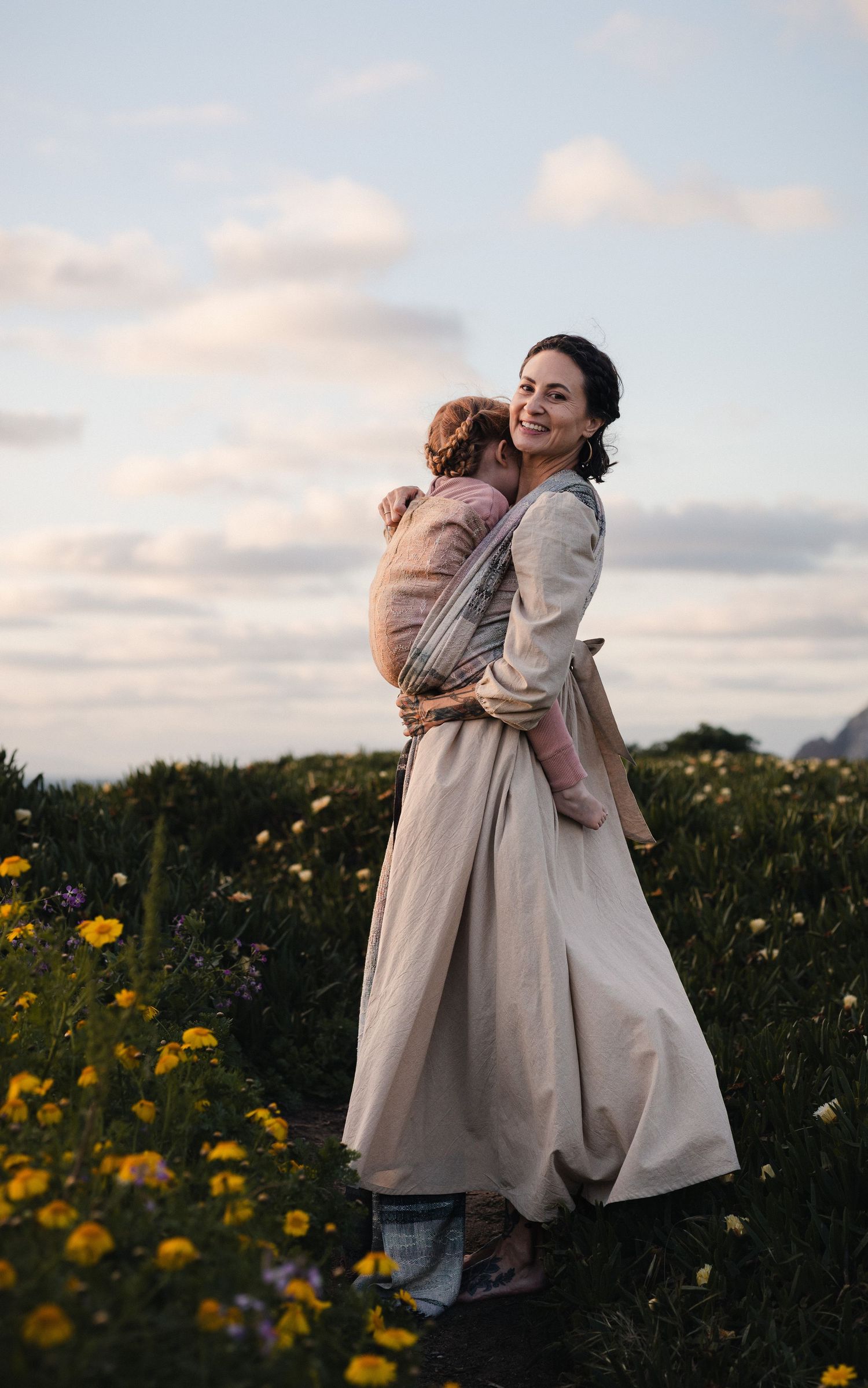 Smiling mother carrying baby in wrap carrier among wildflowers at sunset in a flowing beige dress.