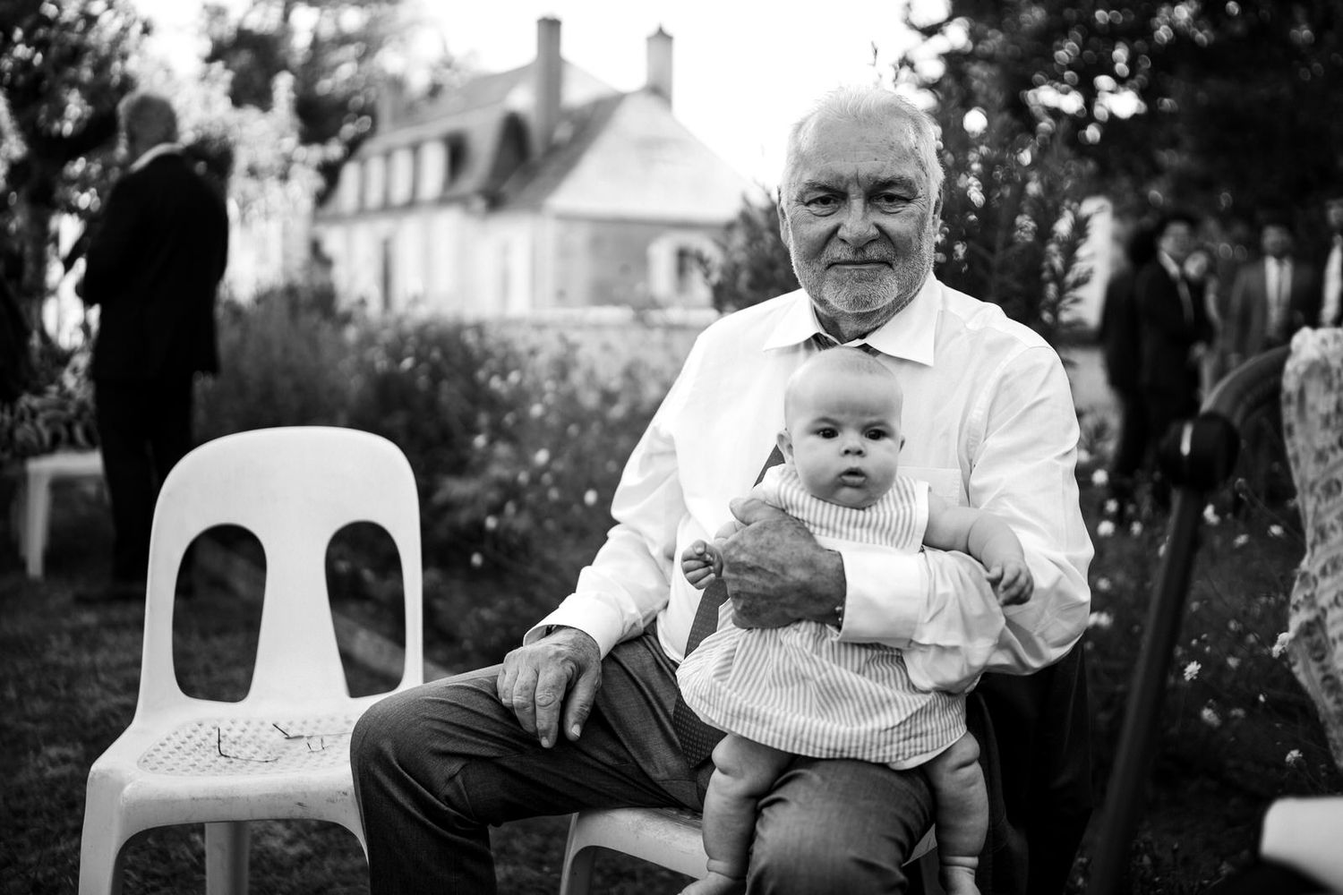 Grand père et petite fille posant pour la photo avec la maison familale en fond, photo en noir et blanc