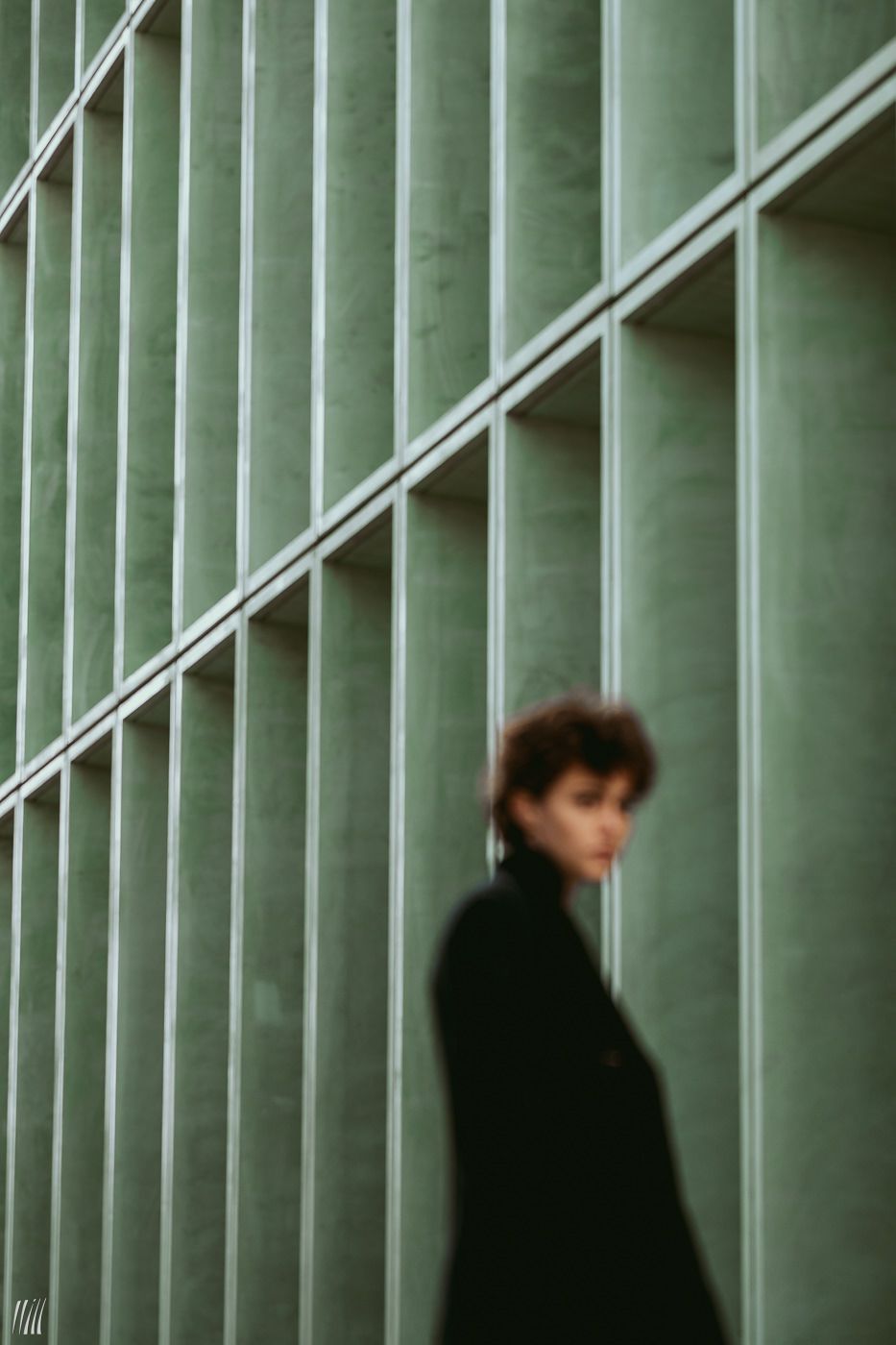 Portrait d'une femme avec un long manteau, devant un bâtiment vert avec une architecture longiligne, le focus est fait sur le bâtiment