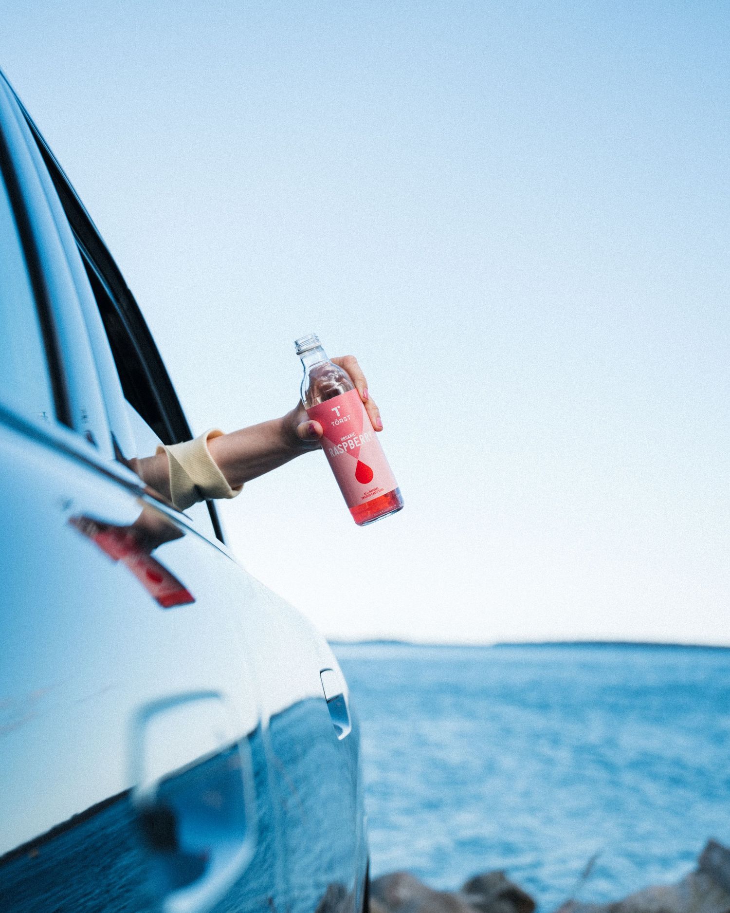 woman holding a bottle of Törst out the window of a car in summer by the ocean. Commercial lifestyle shoot