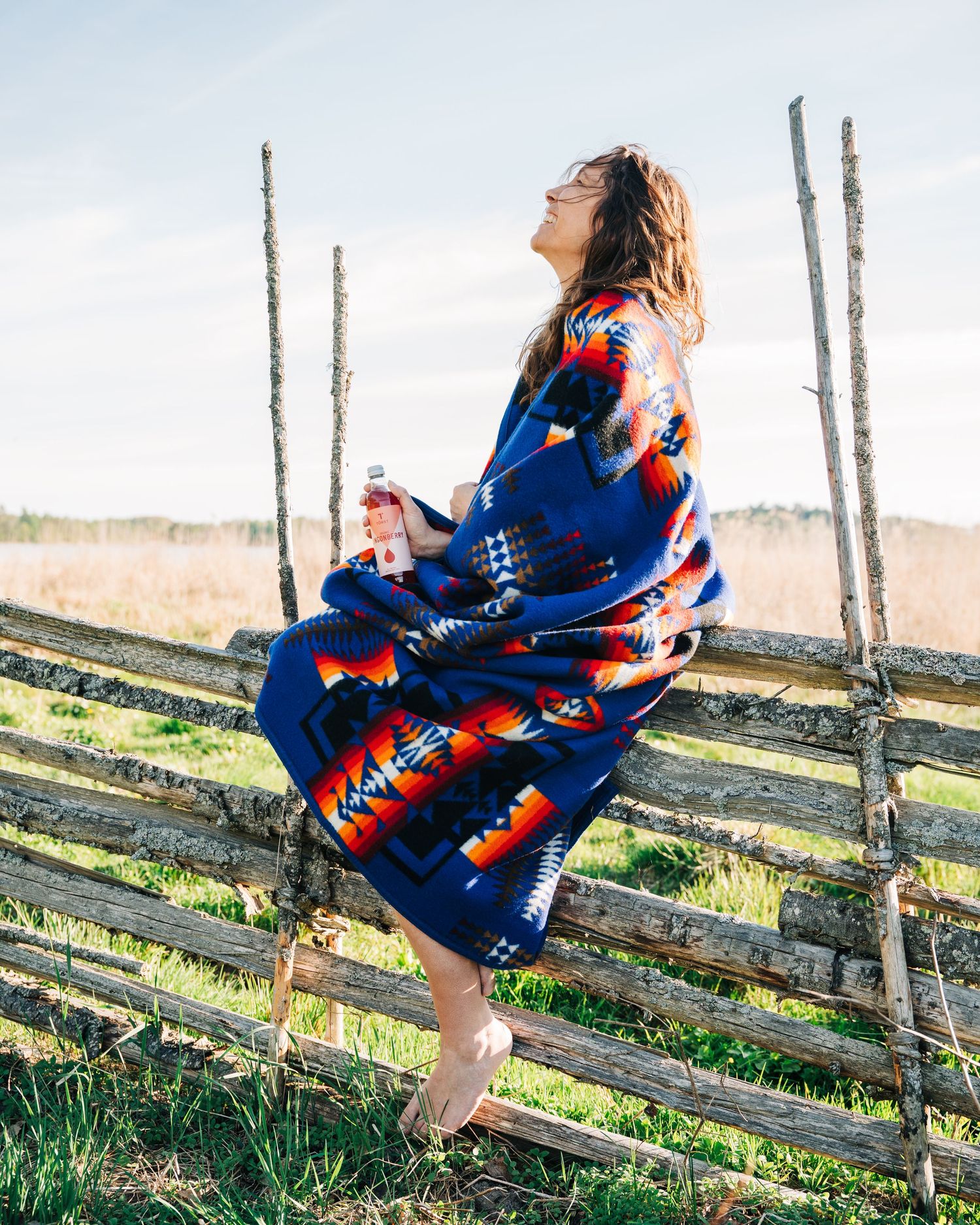 commercial photoshoot of a woman sitting on a fence at sunset drinking Törst lingonberry drink in Sweden