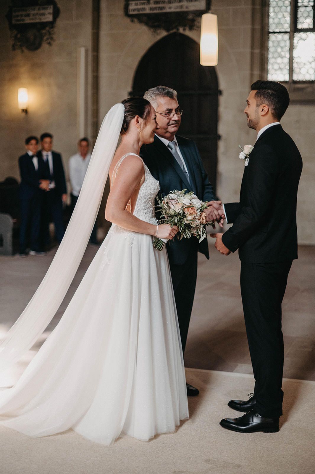 A wedding ceremony takes place in an elegant church interior with a bride in white gown and veil beside officiant and groom.