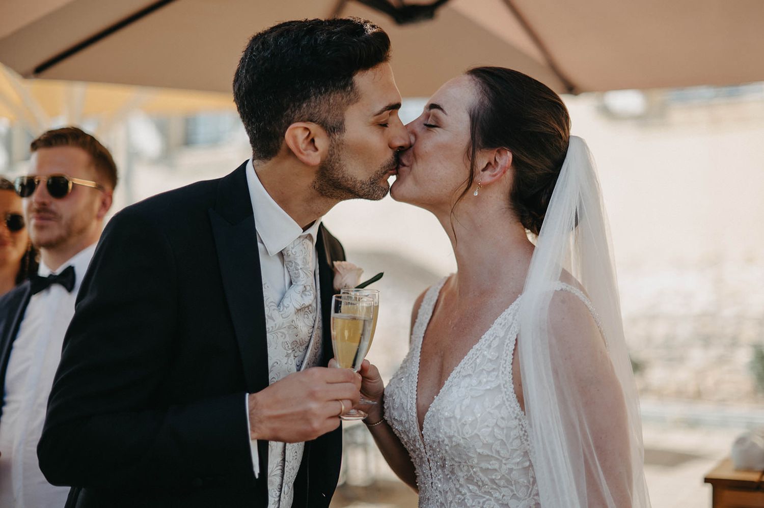 A couple shares a tender kiss during their wedding celebration under a beige tent canopy.